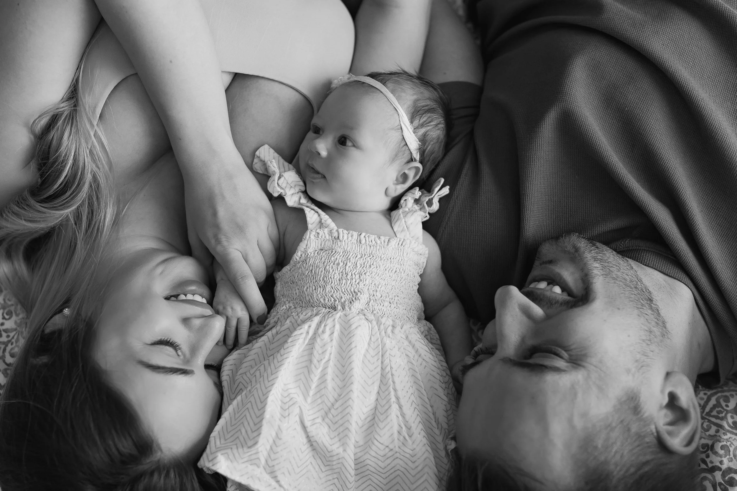 Black-and-white photo of a woman breastfeeding a baby while lying down on a bed, surrounded by four other children. The children are smiling and looking at the baby, who is wearing a headband and a dress.