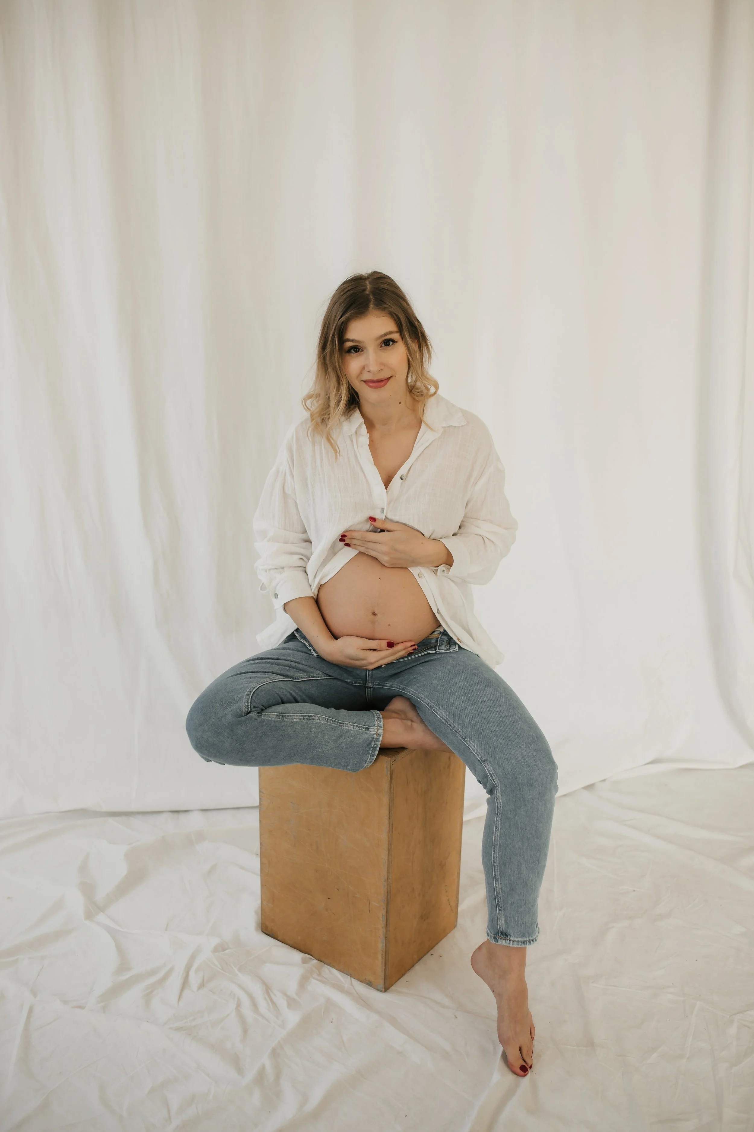 A pregnant woman with light brunette hair and fair skin, sitting cross-legged on a block, holding her belly with one hand, wearing a white shirt and blue jeans, smiling at the camera, in front of a plain white backdrop.