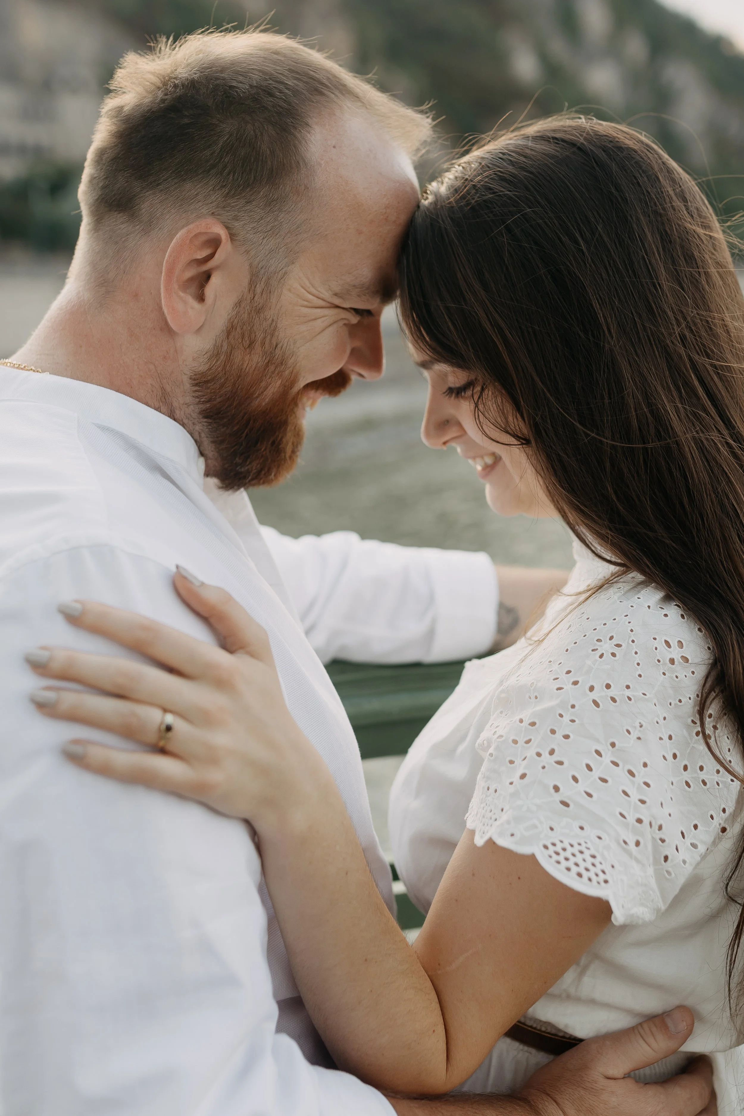A man and woman are embracing each other with their foreheads touching and eyes closed, smiling. The woman has long brown hair and is wearing a white eyelet dress. The man has short light brown hair, a beard, and is wearing a white shirt.