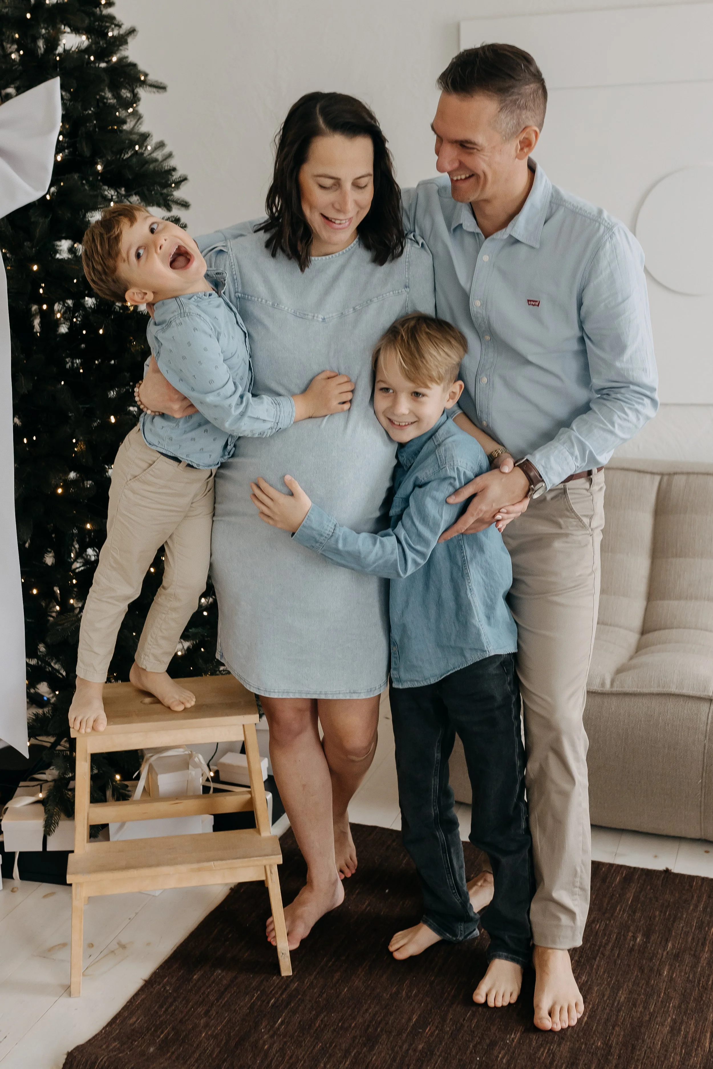 Family celebrating Christmas indoors, with a decorated Christmas tree in the background, embracing each other joyfully, a pregnant woman, two young boys, all smiling.