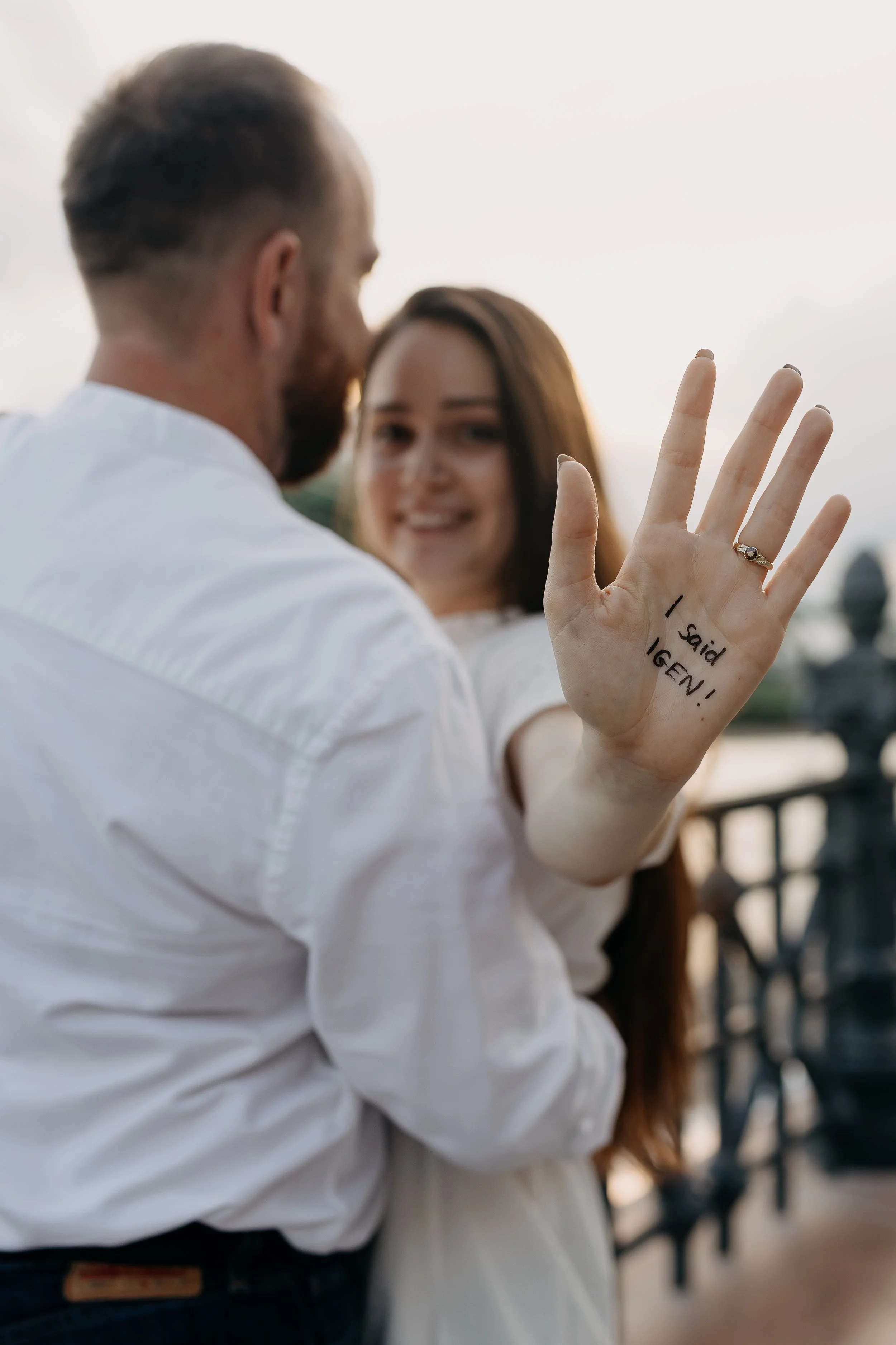 A woman showing her palm with handwritten message, standing next to a man, with a blurred background of outdoors.