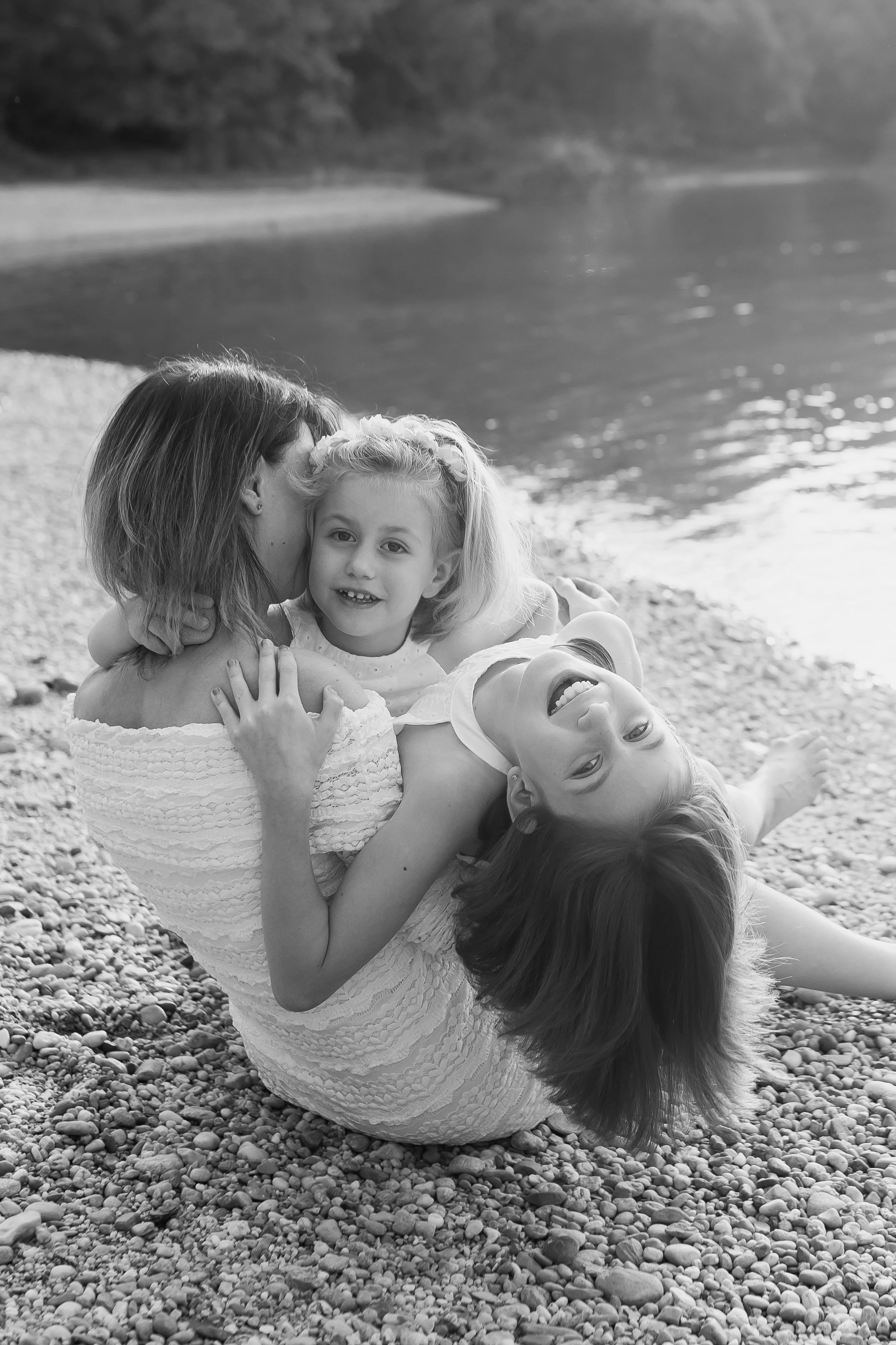 Black and white photograph of four women lying on a pebble beach holding a young girl between them, with a lake and rocky shoreline in the background.