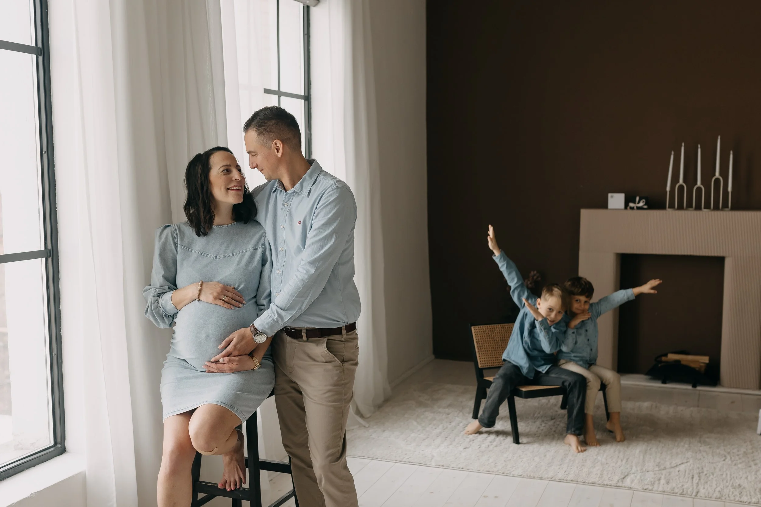 A pregnant woman and a man are standing close, smiling and looking at each other by a large window with white curtains. In the background, three children are sitting on a chair, enthusiastically raising their arms and smiling.