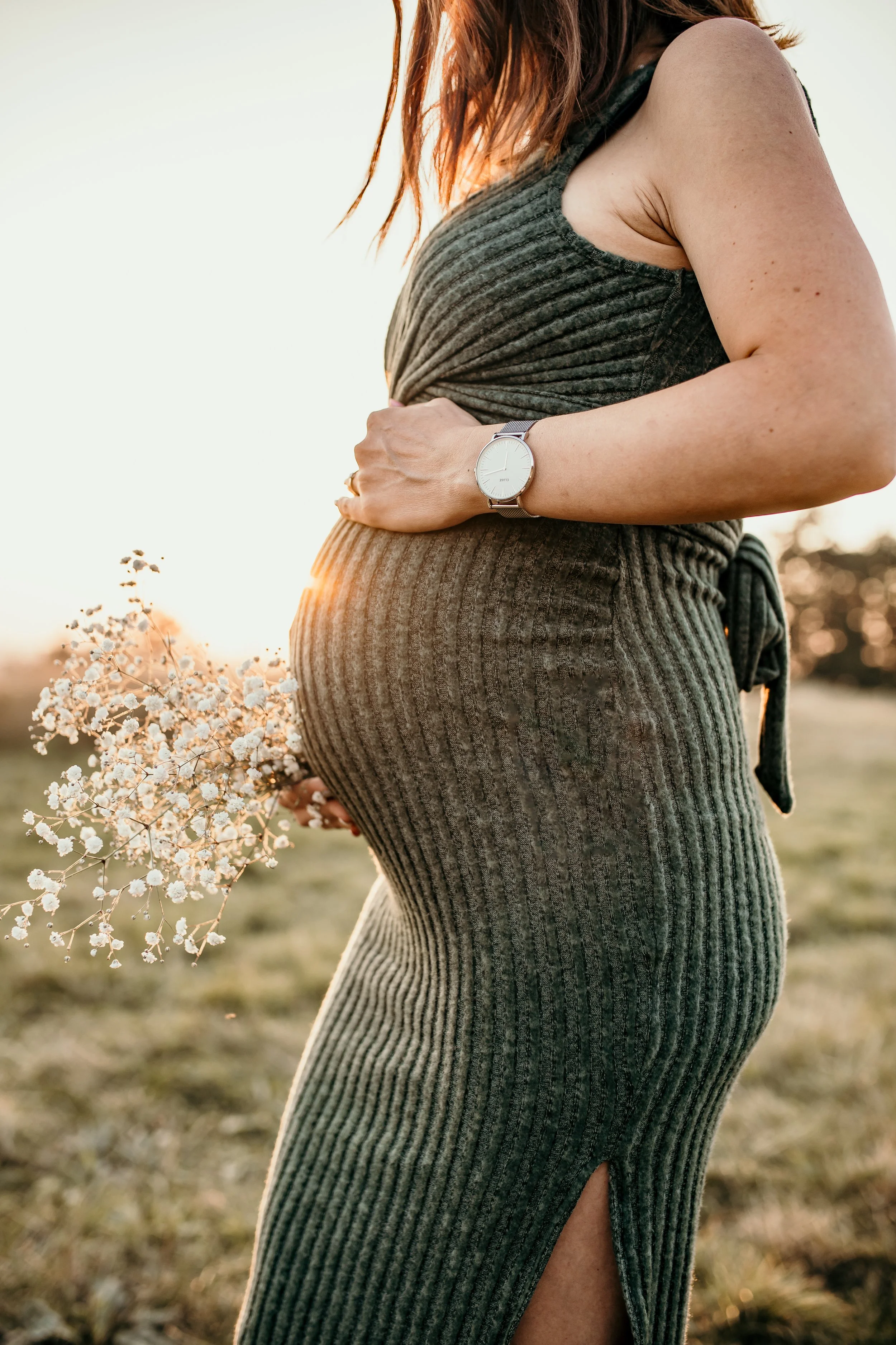 A pregnant woman in a sleeveless ribbed dress holding a small bouquet of white flowers, wearing a watch, outdoors during sunset.