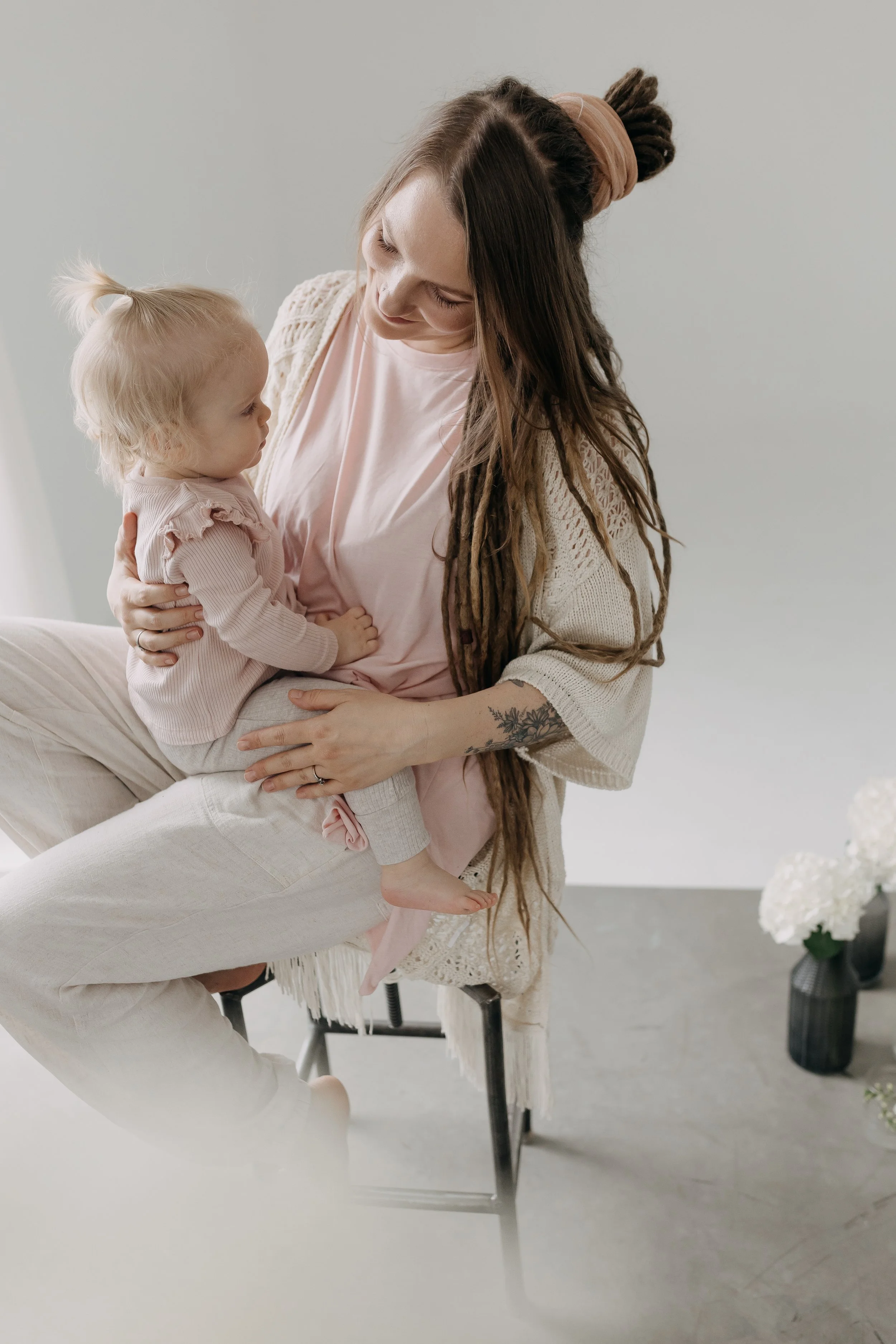 A woman with long brown dreadlocks and a tattoo on her arm holding a young blonde girl with a bow in her hair, sitting on her lap in a bright, minimalistic room.