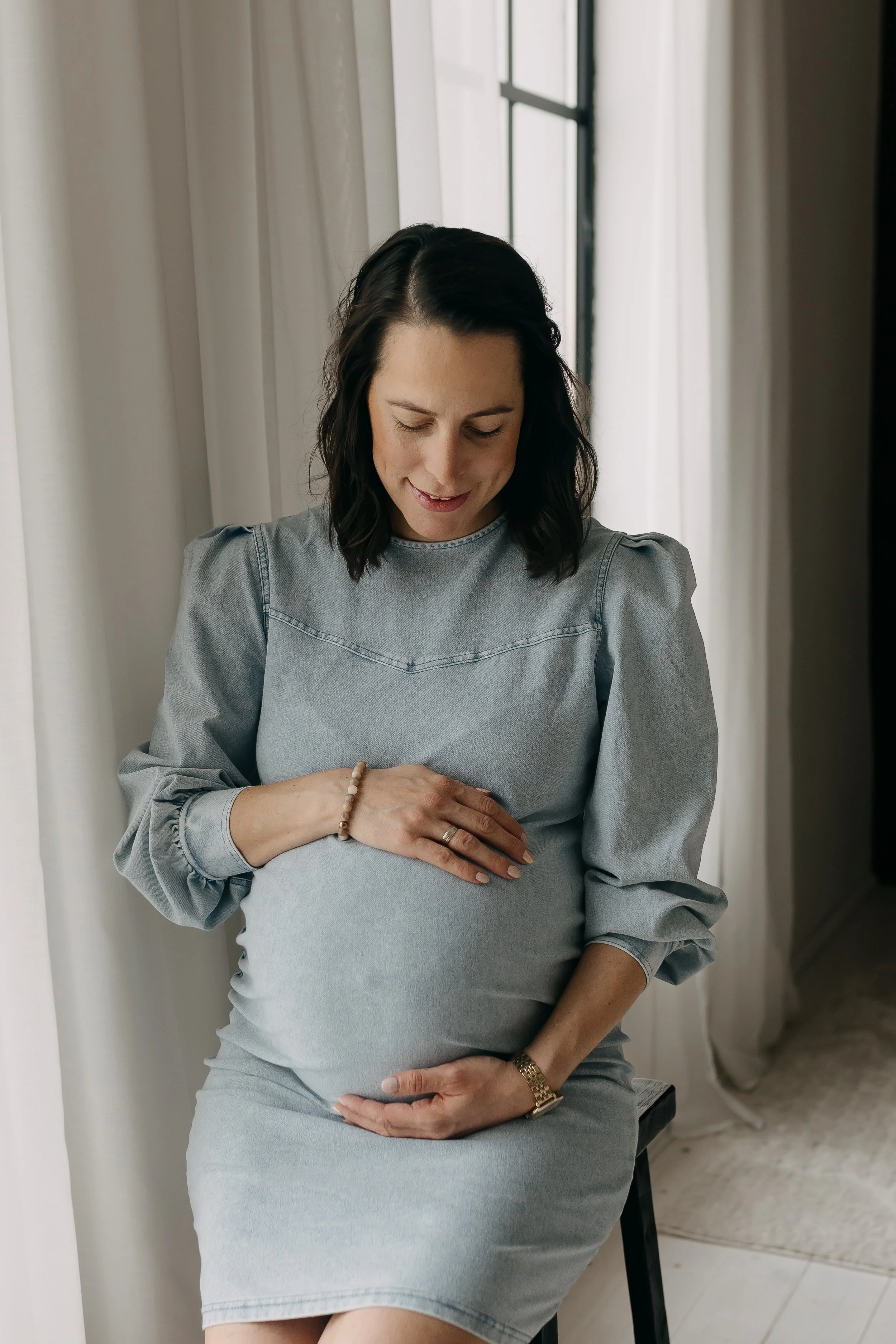 A pregnant woman sitting indoors with her eyes closed, holding her baby bump with both hands, near a window with white curtains.
