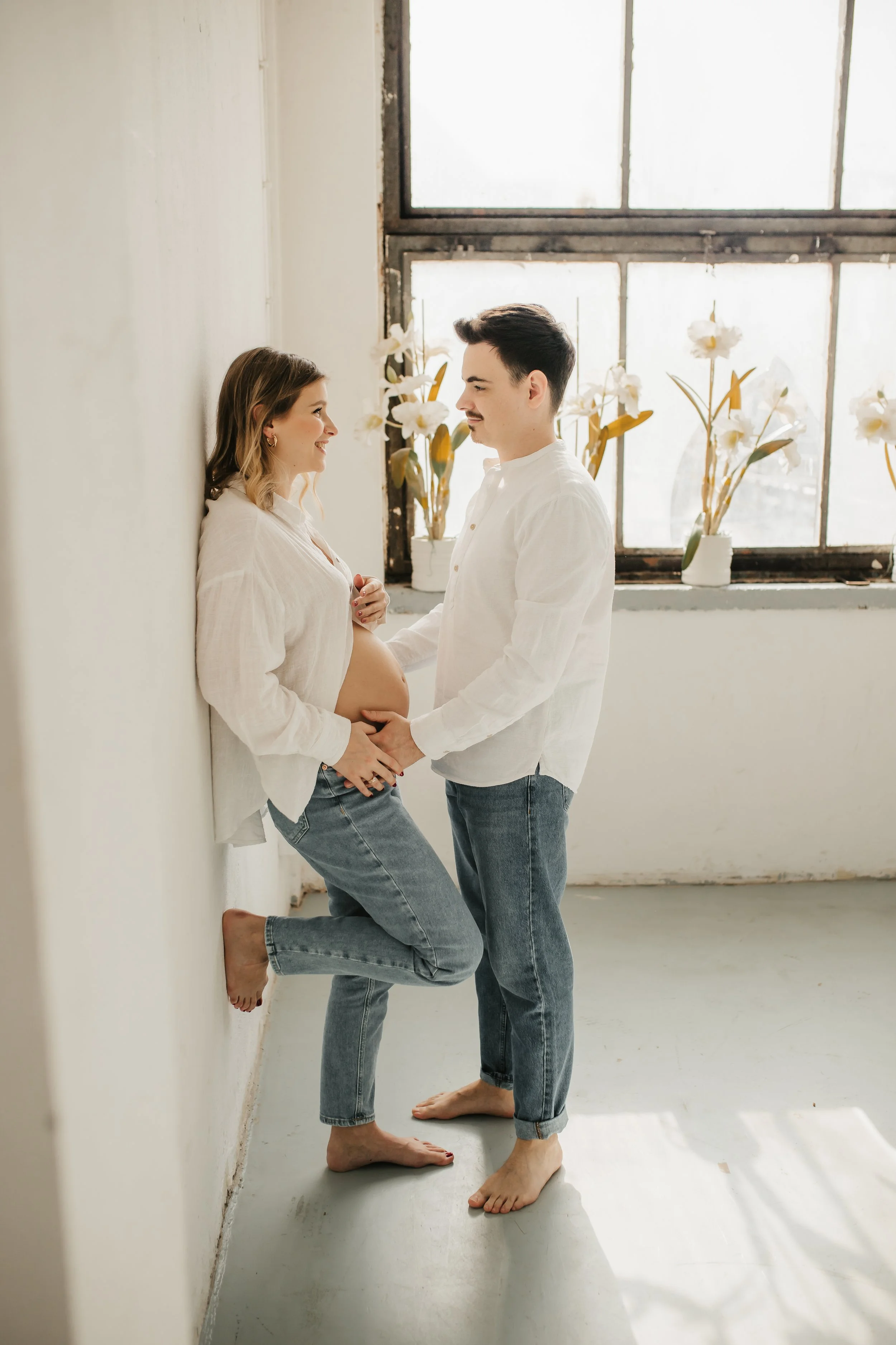 A pregnant woman and a man holding hands face each other in a bright room with large windows and potted white flowers in the background. The woman is leaning against a white wall, and the man is standing barefoot on a white floor.