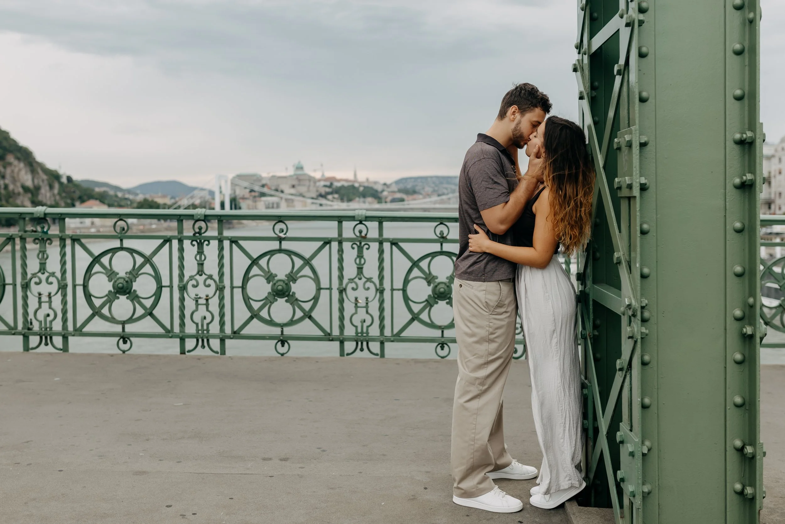 A couple sharing a kiss on a bridge with a cityscape in the background, including a bridge and buildings under a cloudy sky.