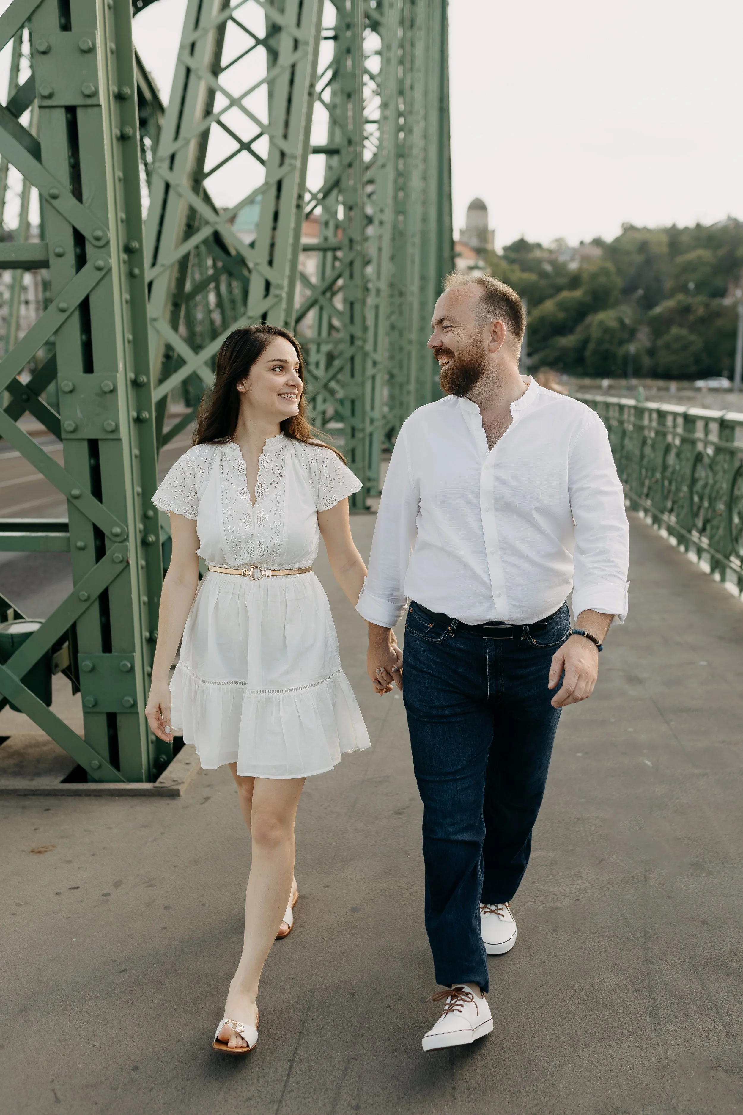 A young woman and a man holding hands and walking together on a bridge, smiling and looking at each other. The woman is wearing a white dress and sandals, and the man is dressed in a white shirt, dark jeans, and white sneakers. The bridge is green wi