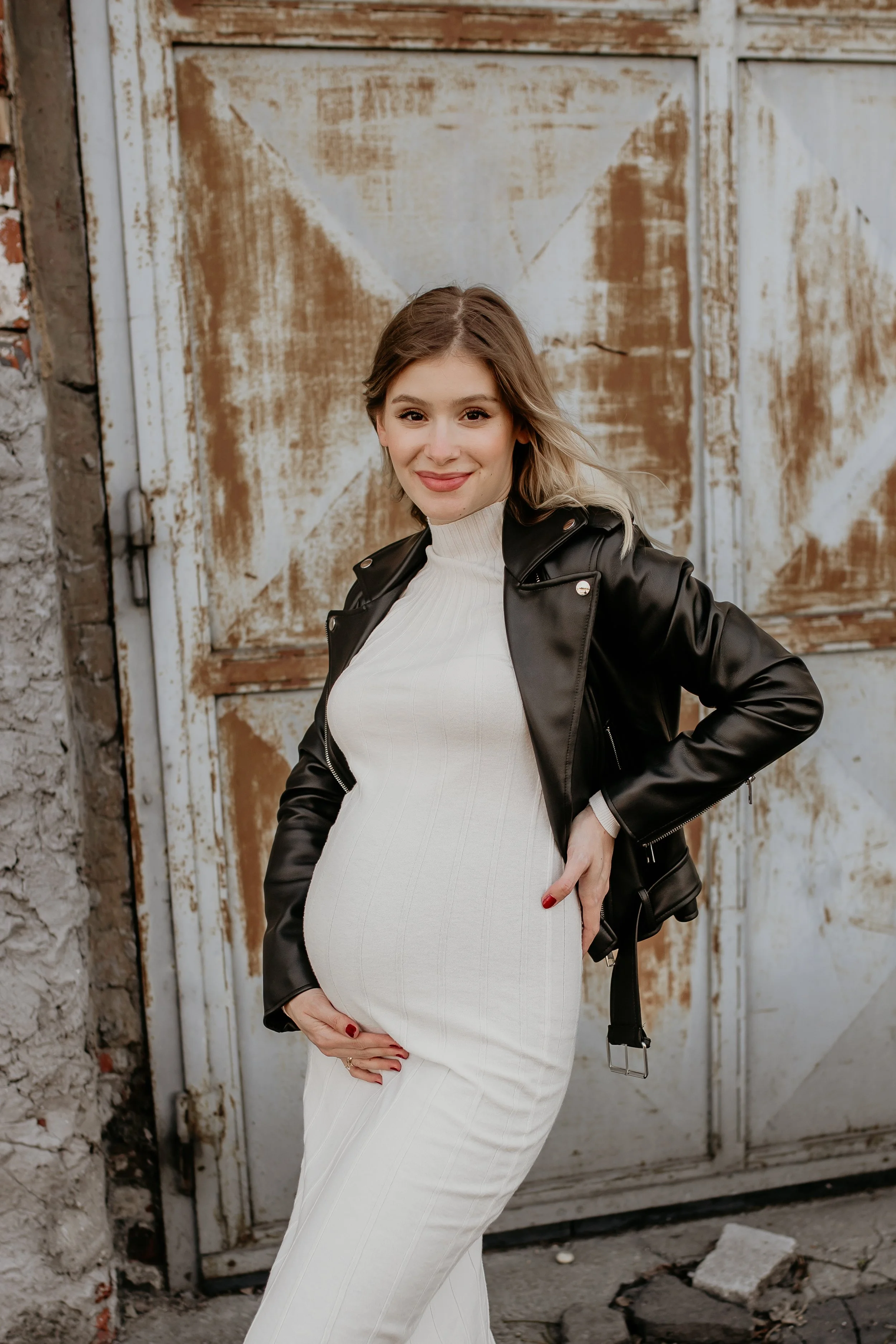 A pregnant woman with blonde hair, wearing a white dress and a black leather jacket, standing outdoors in front of a weathered metal door, smiling and holding her belly.