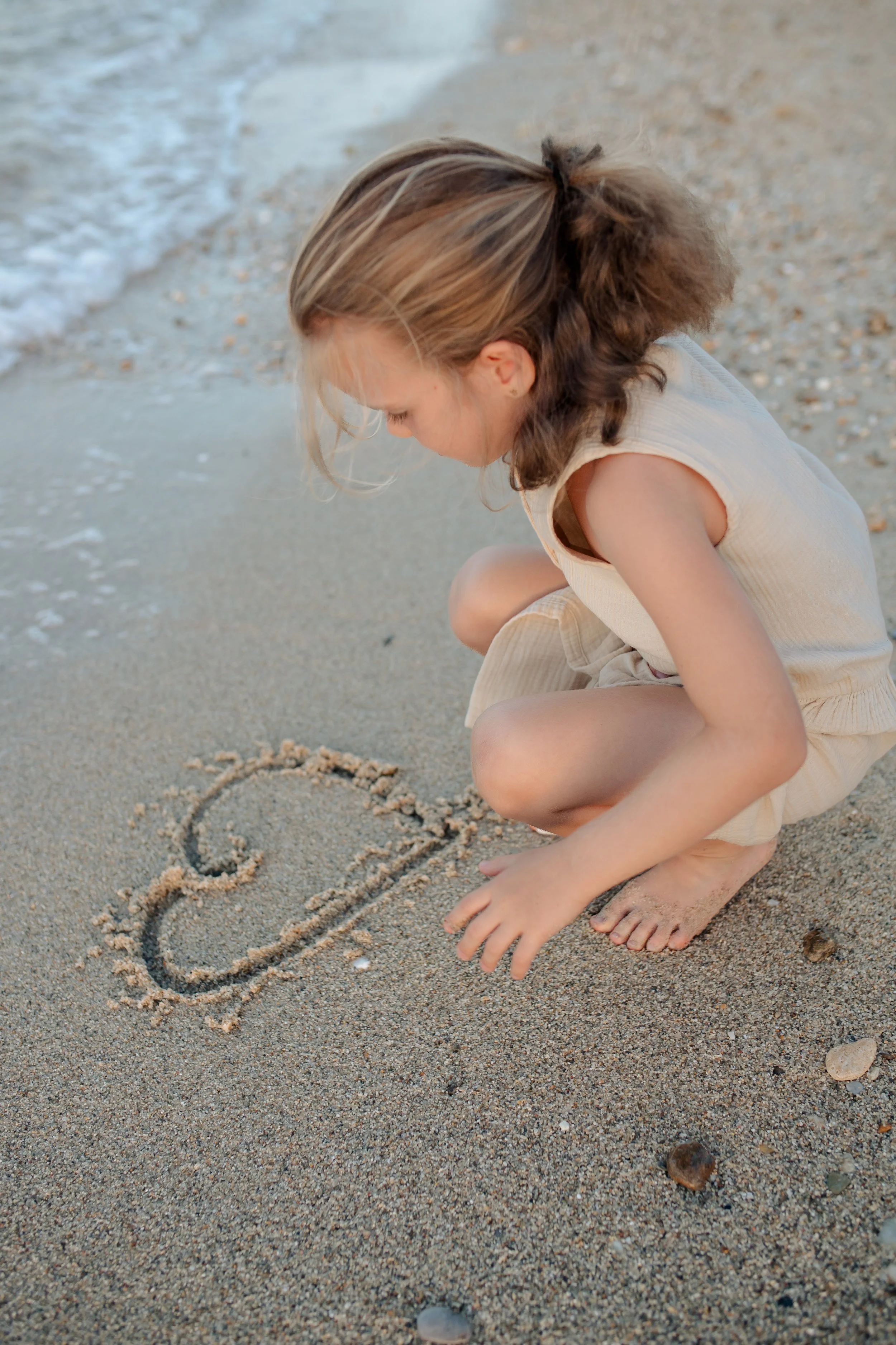 A young girl with light brown hair and wearing a sleeveless beige dress, squatting on a sandy beach, drawing a heart in the sand with her finger.