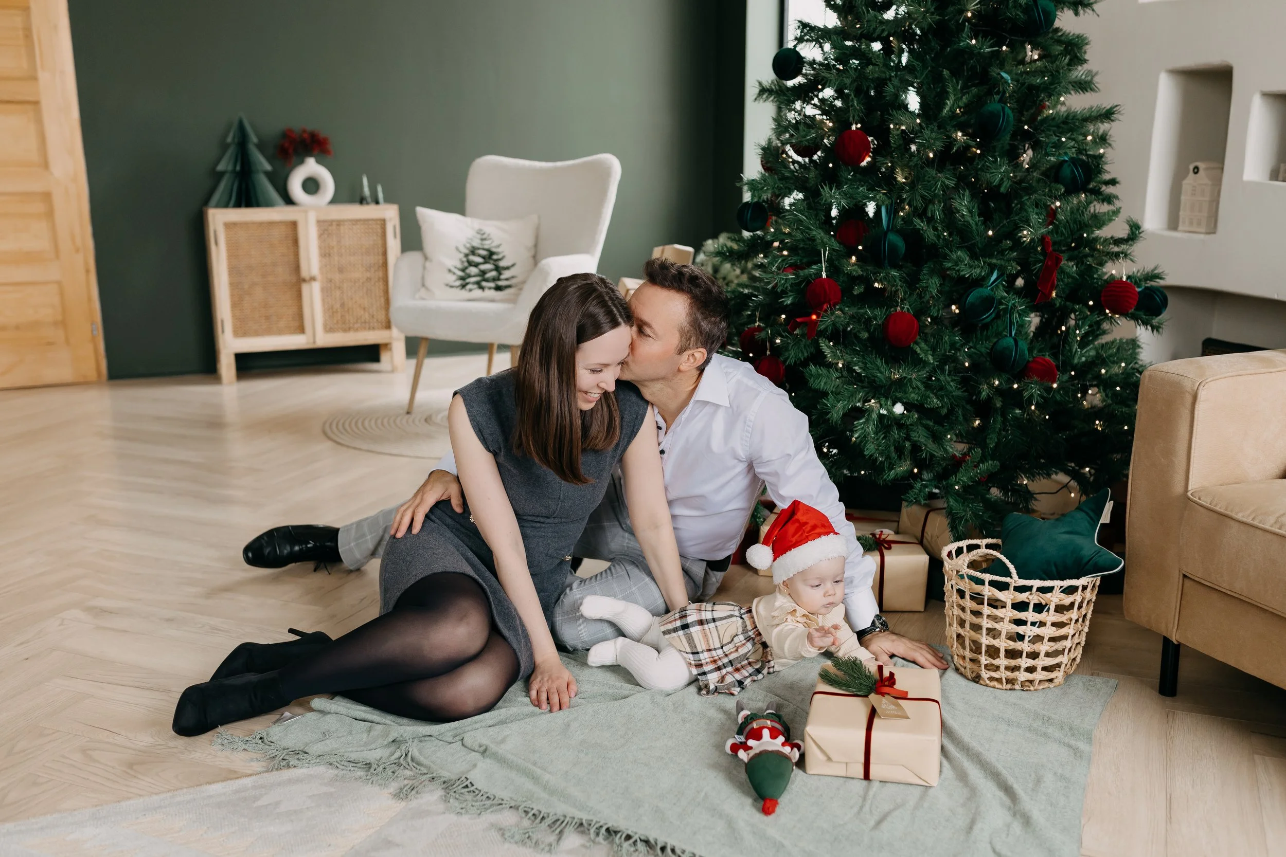 A family celebrating Christmas with a decorated tree, presents, and a cozy living room setting. A man and woman sit on the floor embracing, with two children nearby, one wearing a Santa hat.