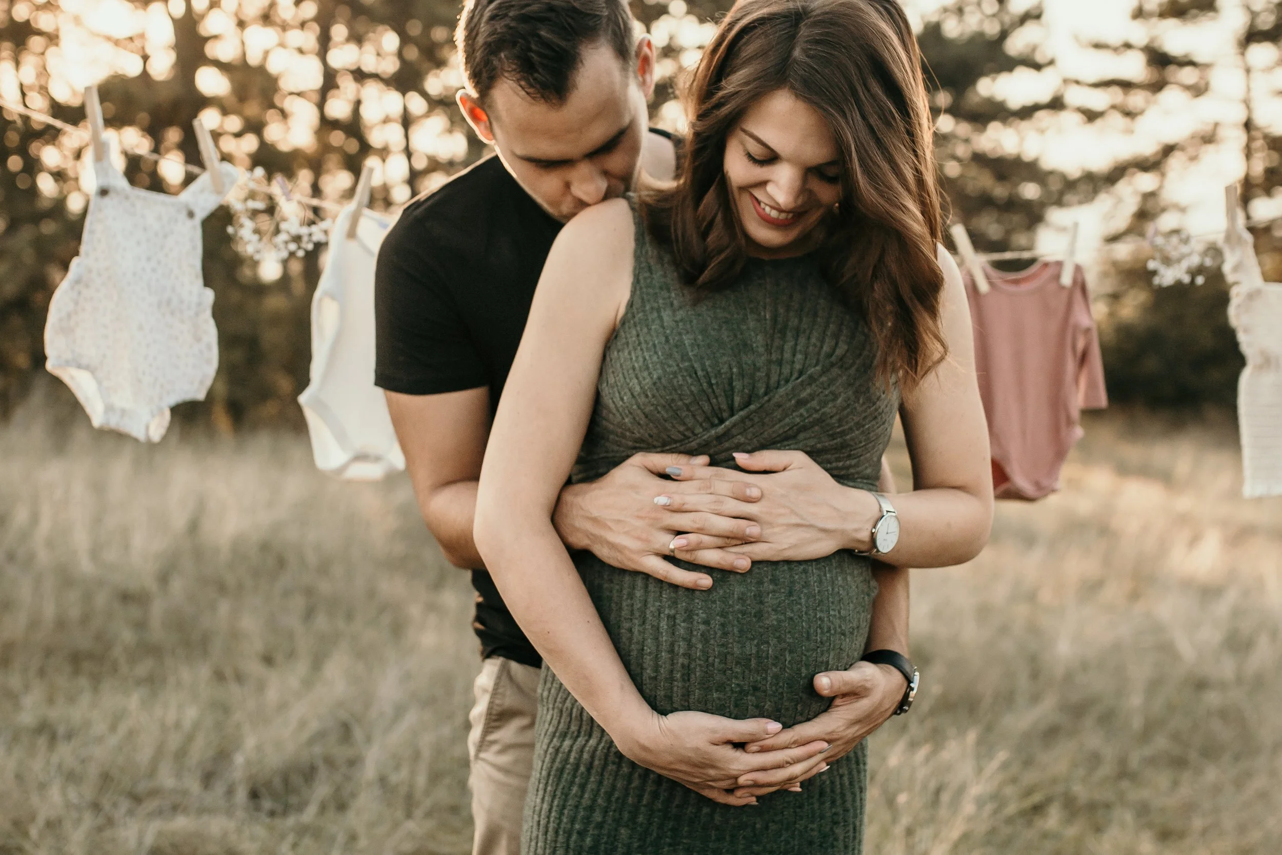 A couple stands outdoors during sunset, embracing with the man behind the woman, both smiling and looking down at her pregnancy belly. In the background, a clothesline with baby clothes hangs among trees.