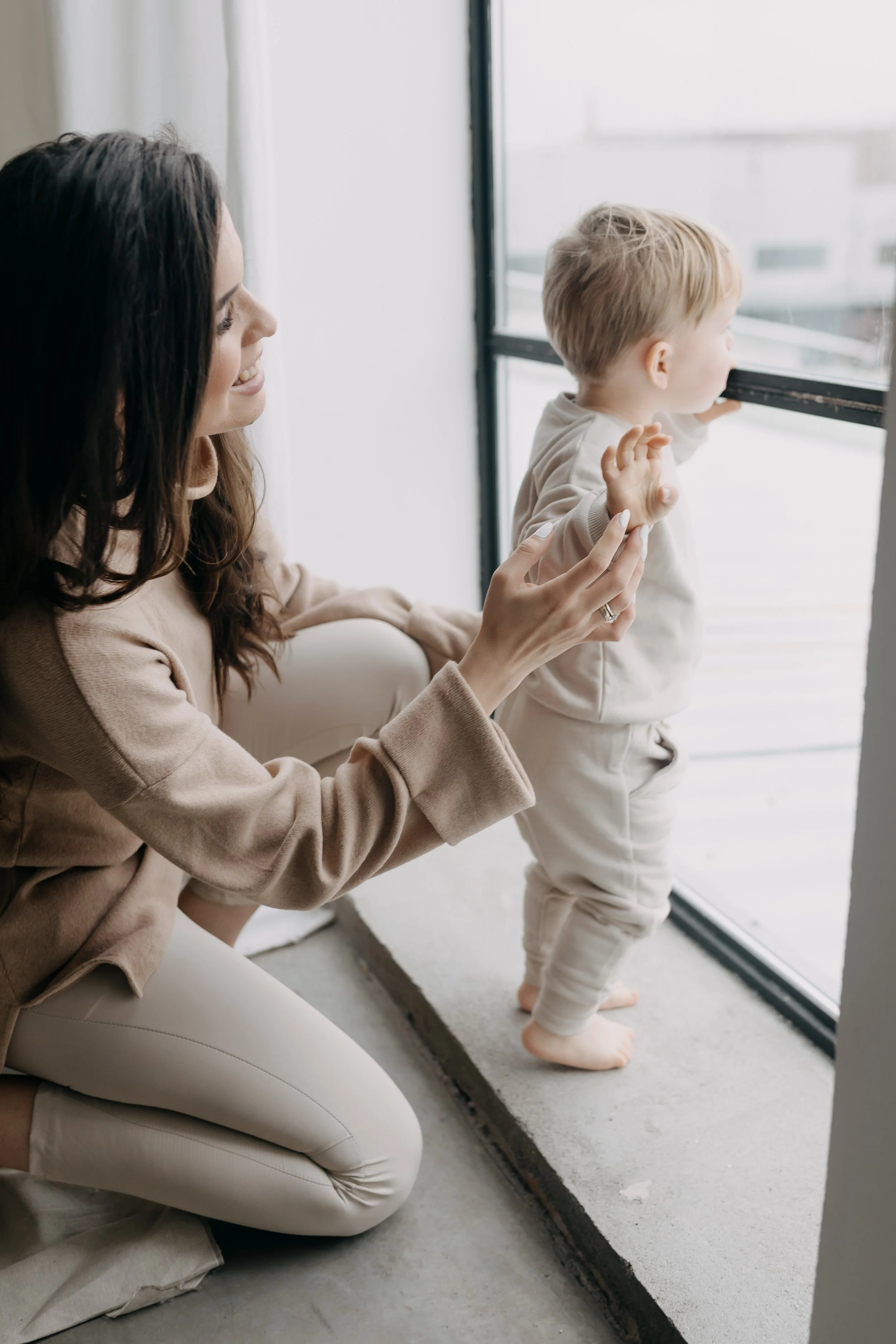 A woman with dark hair and beige clothing smiling while holding hands with a blonde-haired toddler looking out a window.