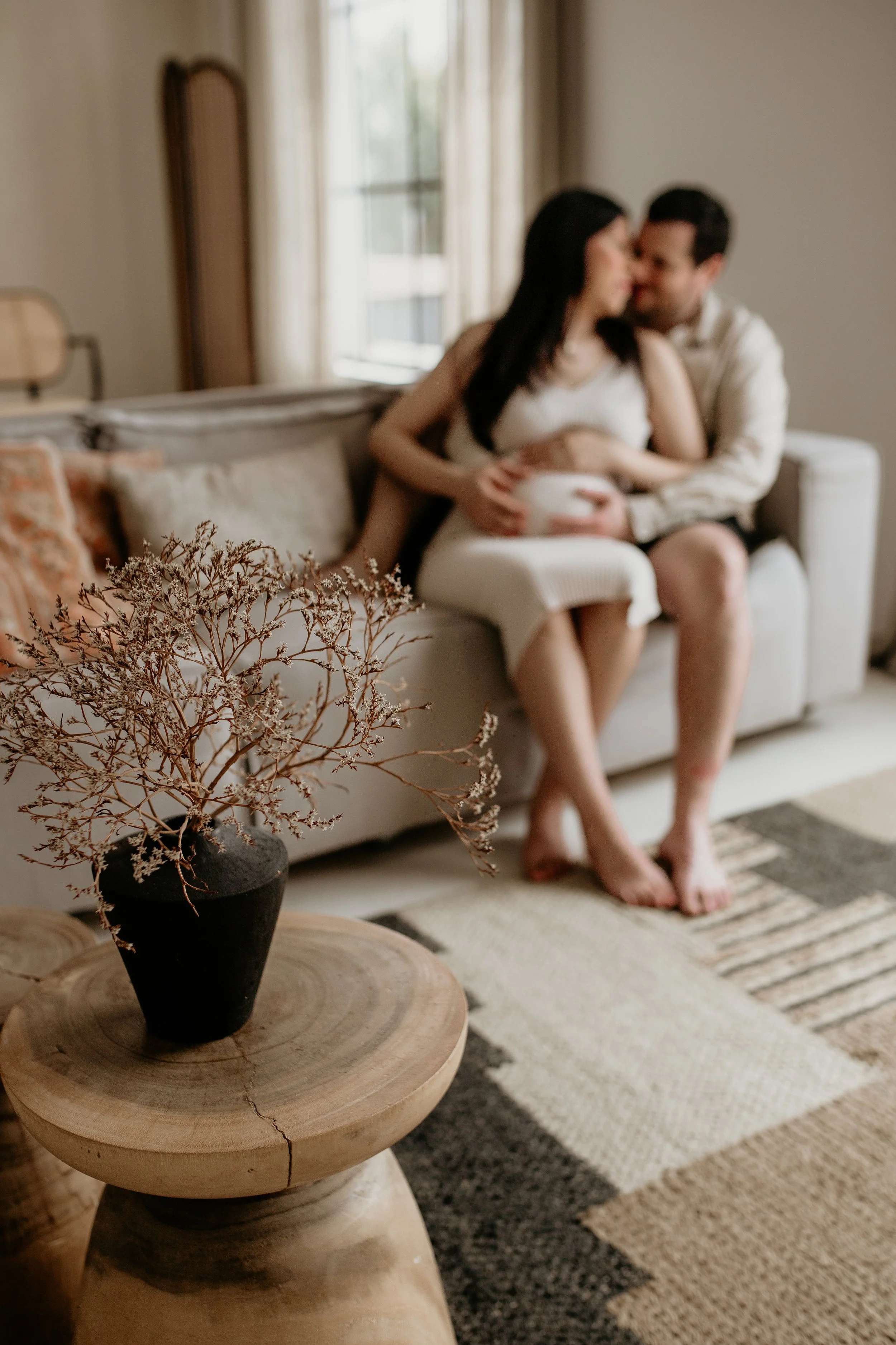 A couple sitting on a sofa in a cozy, well-lit living room, with a wooden coffee table holding a black vase with dried flowers in the foreground.