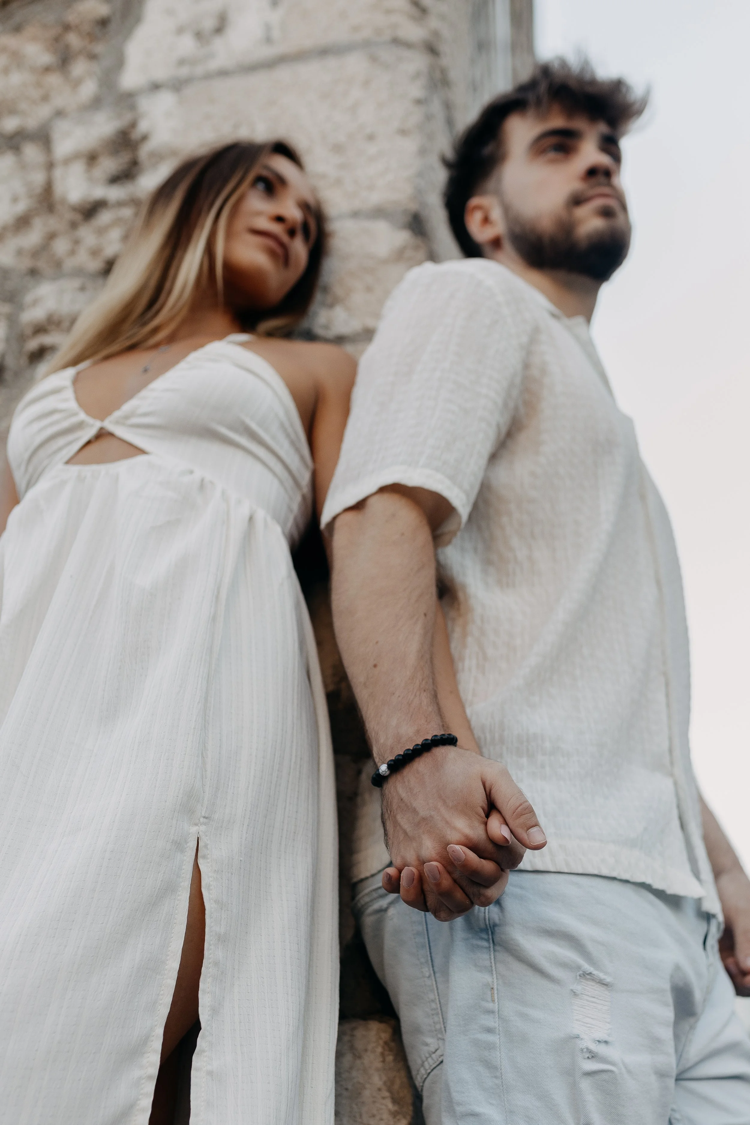 A couple holding hands, standing outdoors near a stone wall, with the woman in a white dress and the man in a white shirt and light jeans.