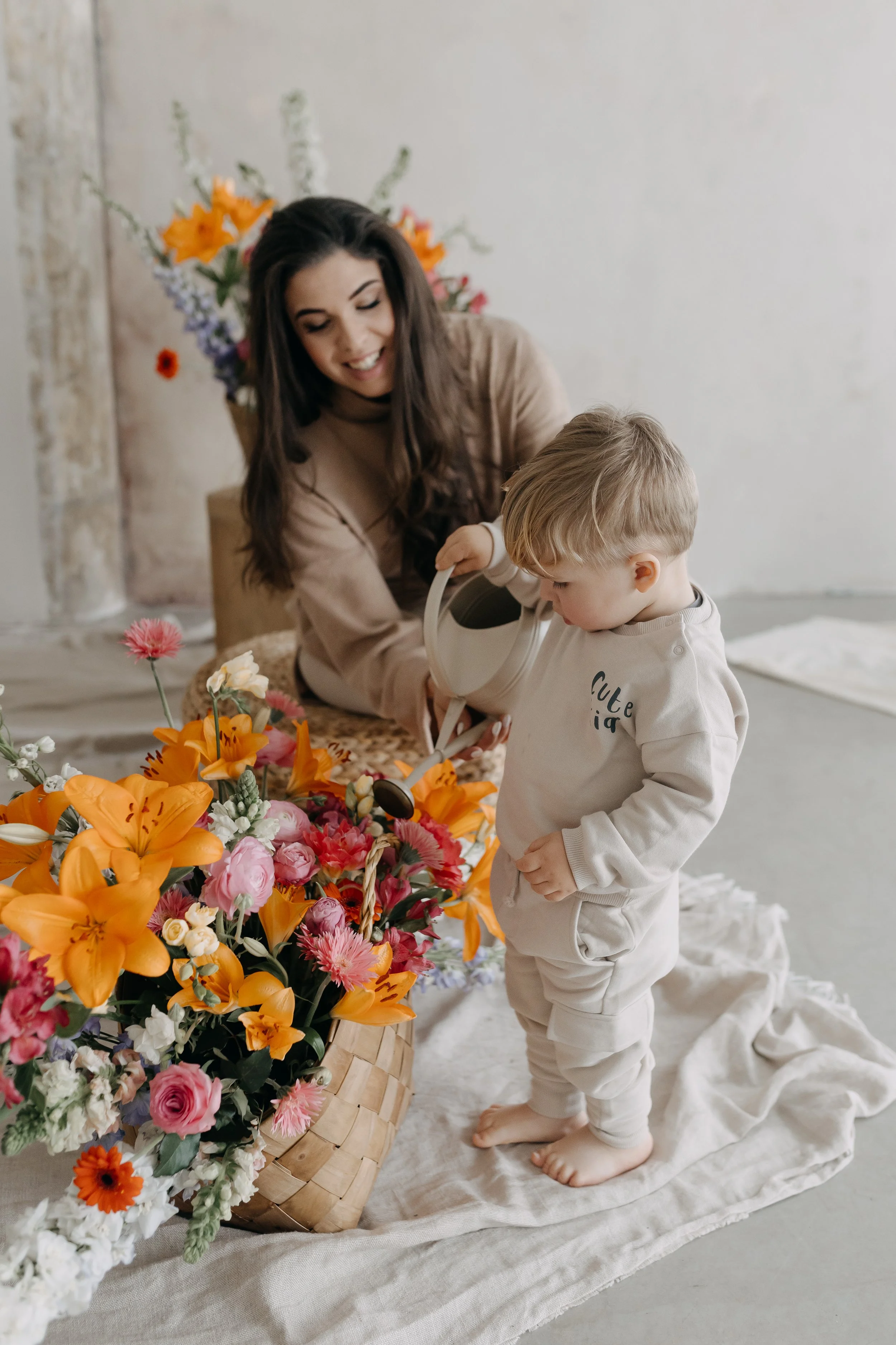 A woman and a young child with blond hair are arranging a large basket of colorful flowers, including orange lilies, pink roses, and other mixed blooms. The woman has long dark hair and is smiling, while the child is focused on watering the flowers w