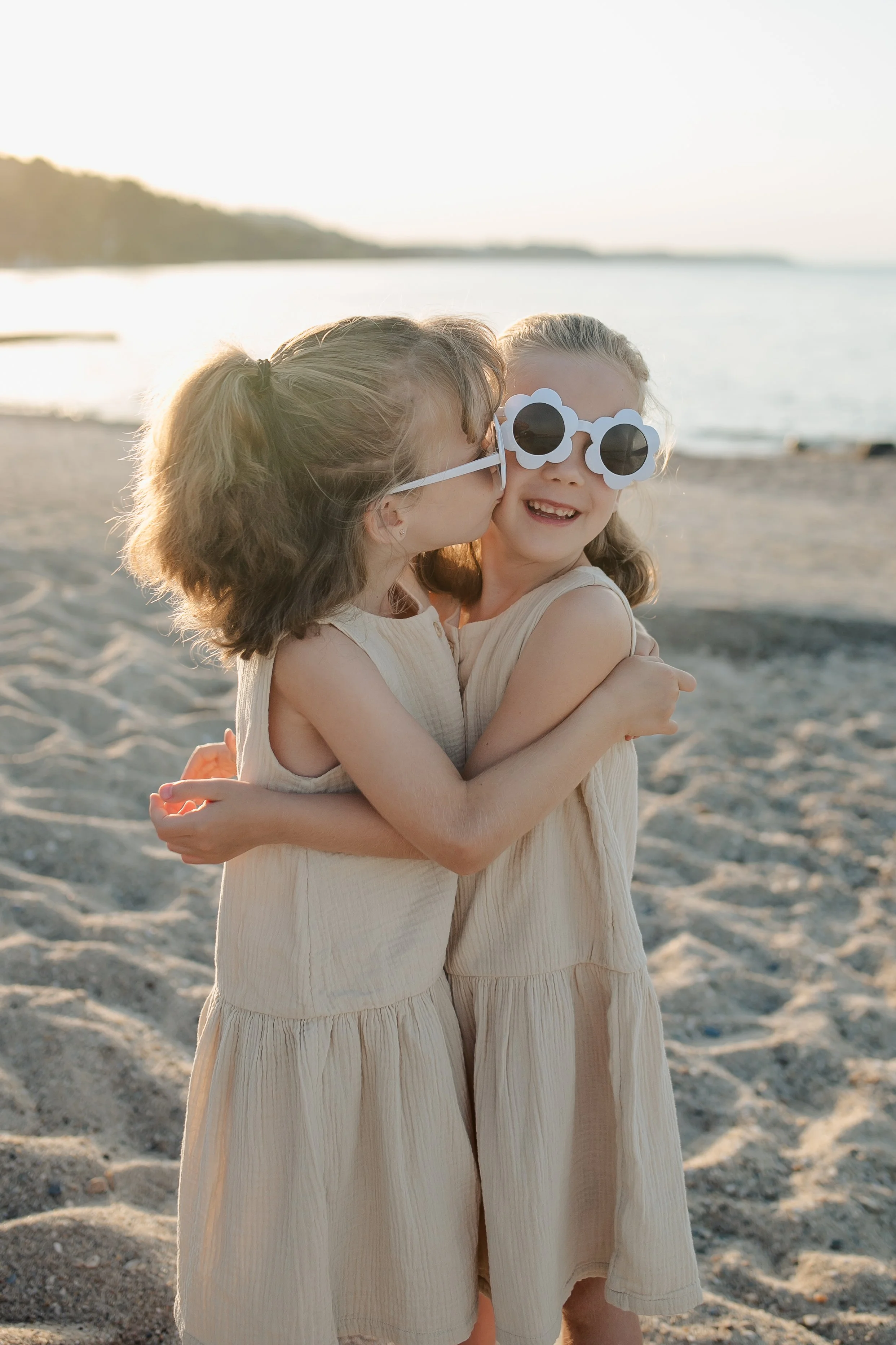 Two young girls hugging on the beach at sunset, wearing matching cream-colored dresses and novelty sunglasses, with a calm body of water and distant land in the background.