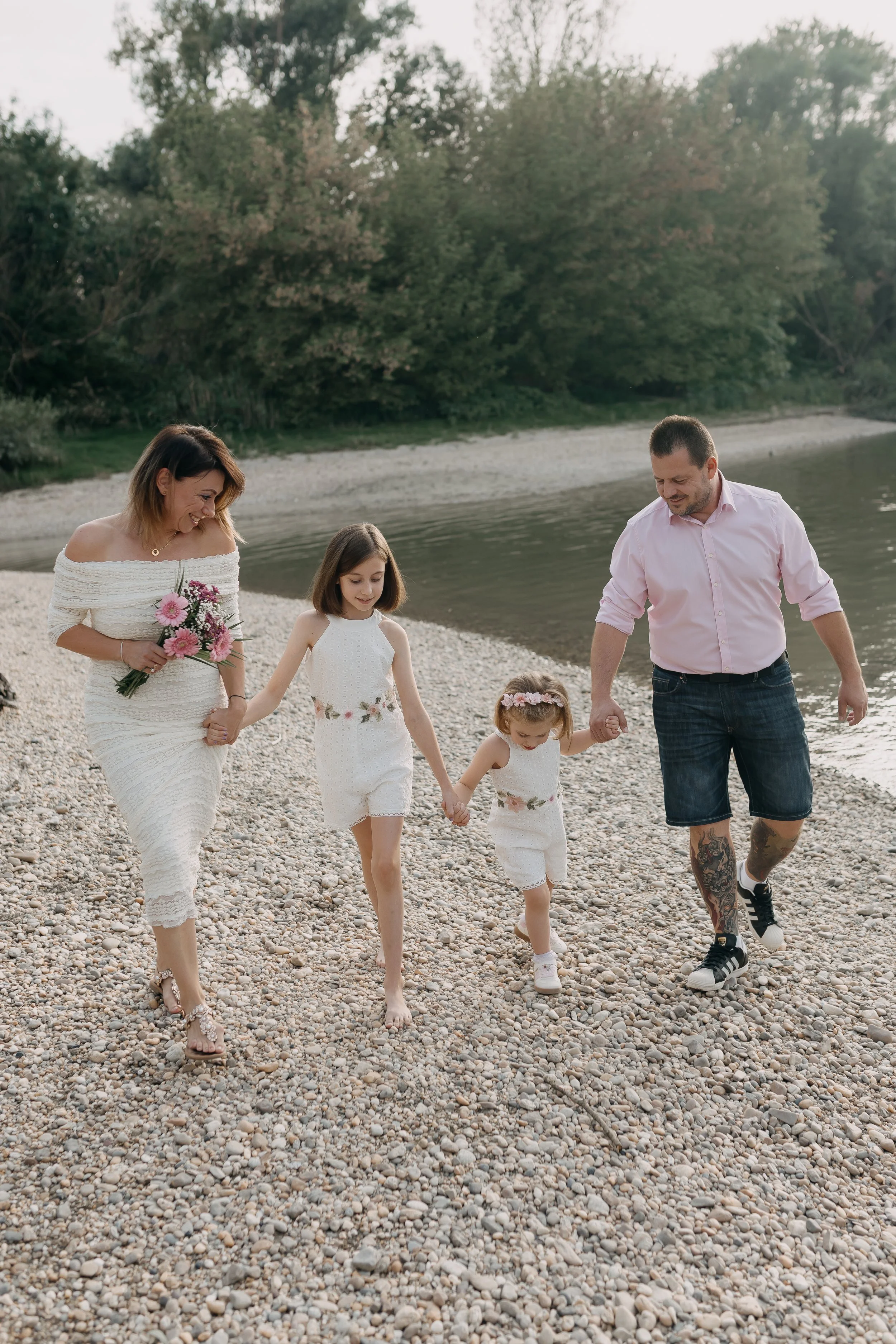 A family of five walking hand in hand along a pebble beach by a river, with green trees in the background.