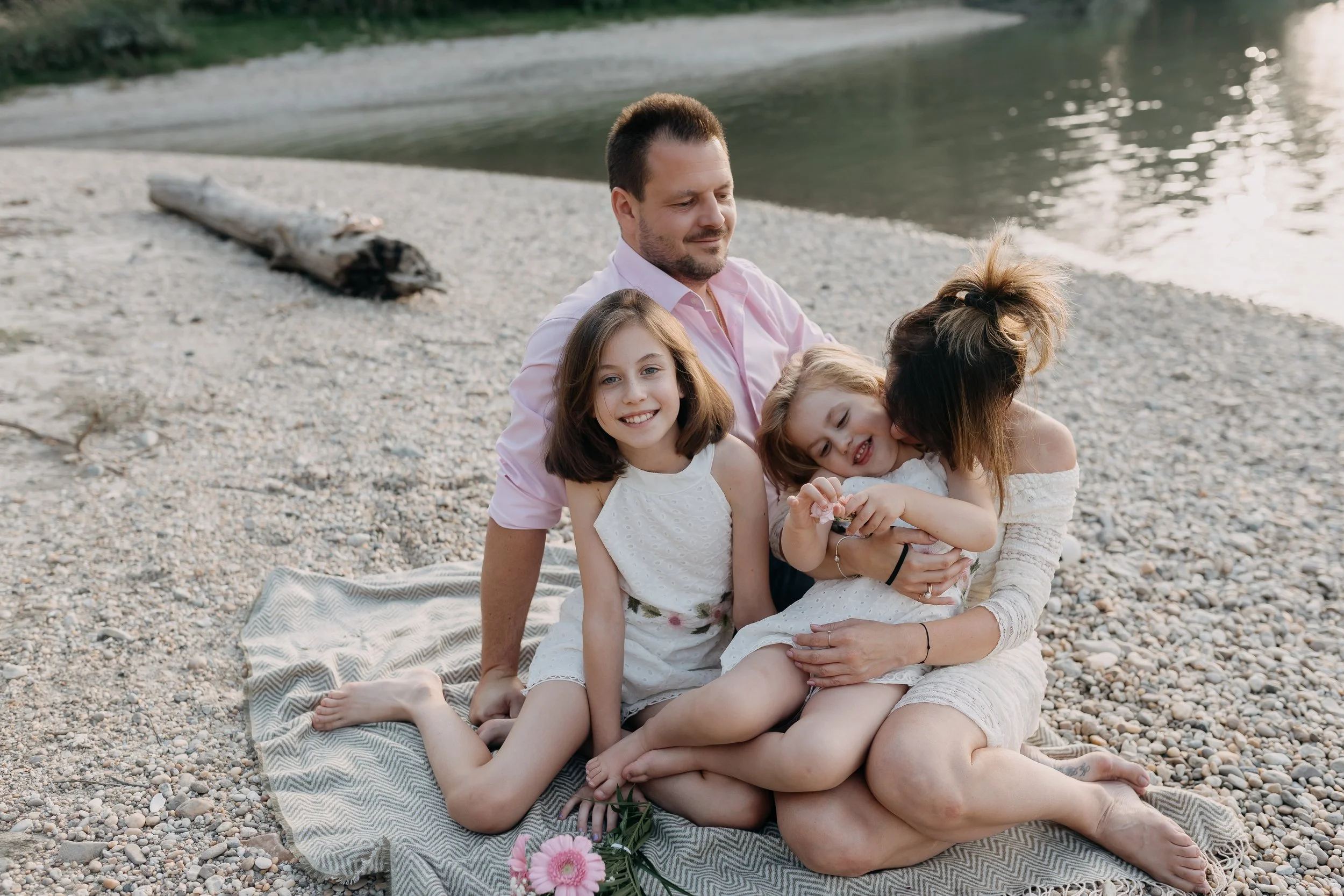 A family sitting on a blanket by the water's edge on a rocky beach, with a log and trees in the background. The mother and three young girls are smiling and cuddling, with pink flowers in the foreground.