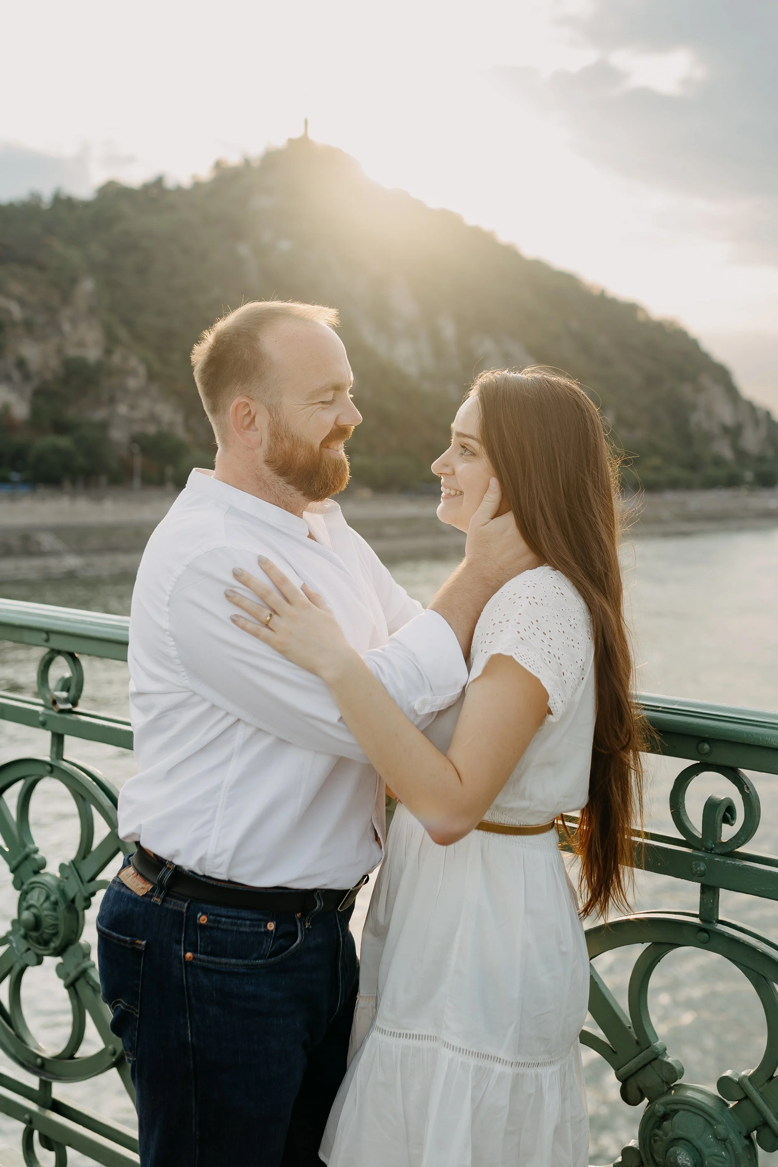 A man with a beard and a woman with long brown hair, smiling and looking at each other, standing on a bridge with a scenic landscape of trees, a hill, and a river in the background. The woman is gently touching the man's face, and they are both weari