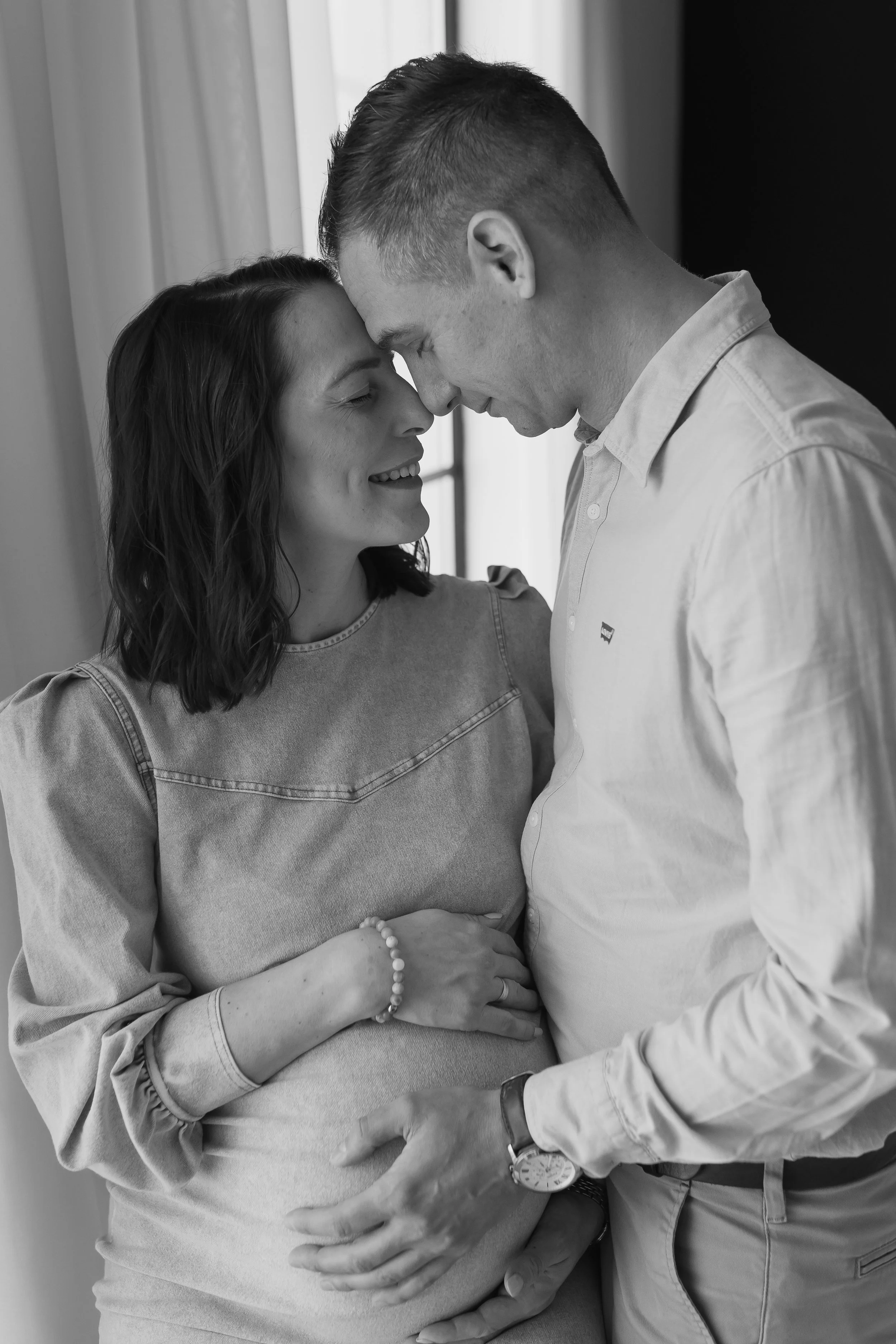 A pregnant woman and a man are touching their foreheads and smiling at each other, with the woman's hand resting on her belly.