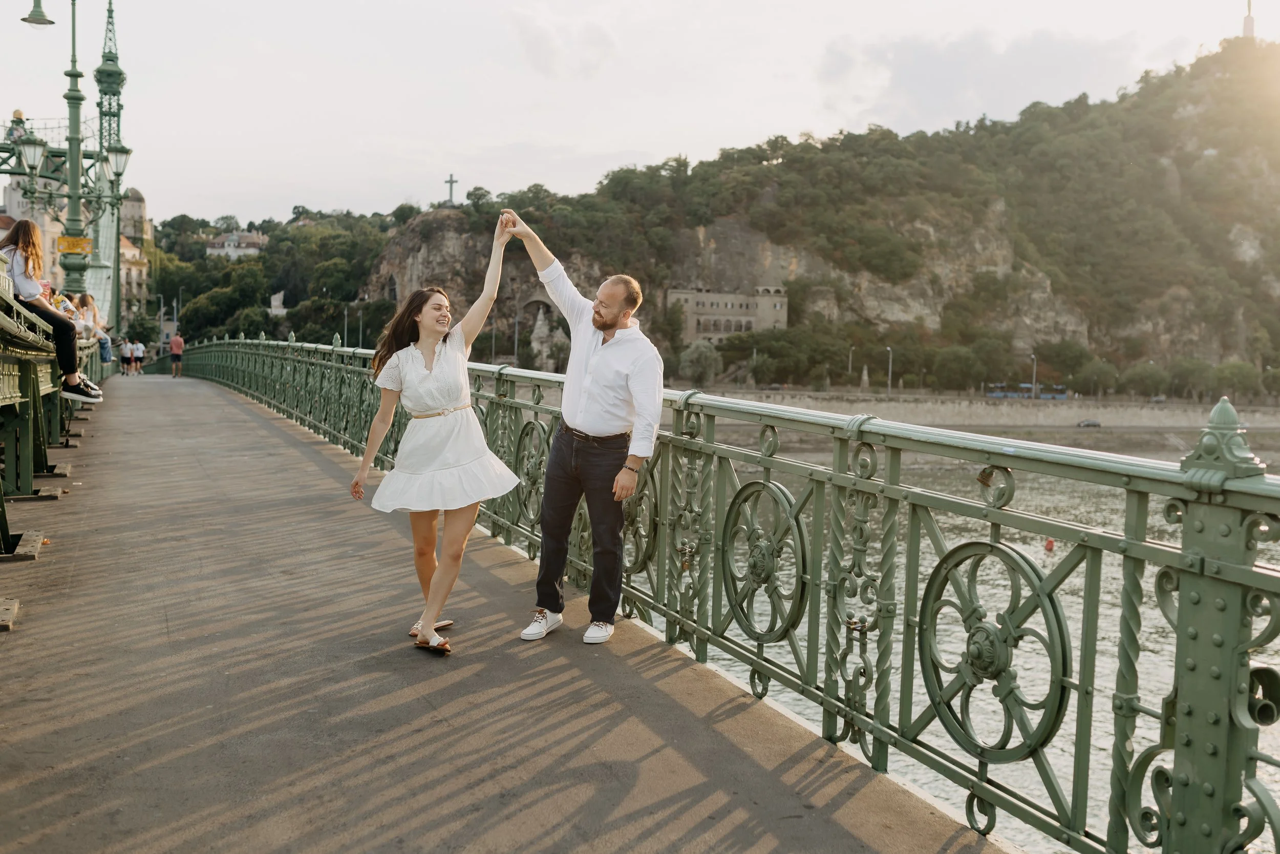 A couple dancing on a bridge in a city park during late afternoon. The woman is wearing a white dress, and the man is dressed casually in a white shirt and dark pants. The background includes a hill with a cross, some buildings, and a decorative ligh