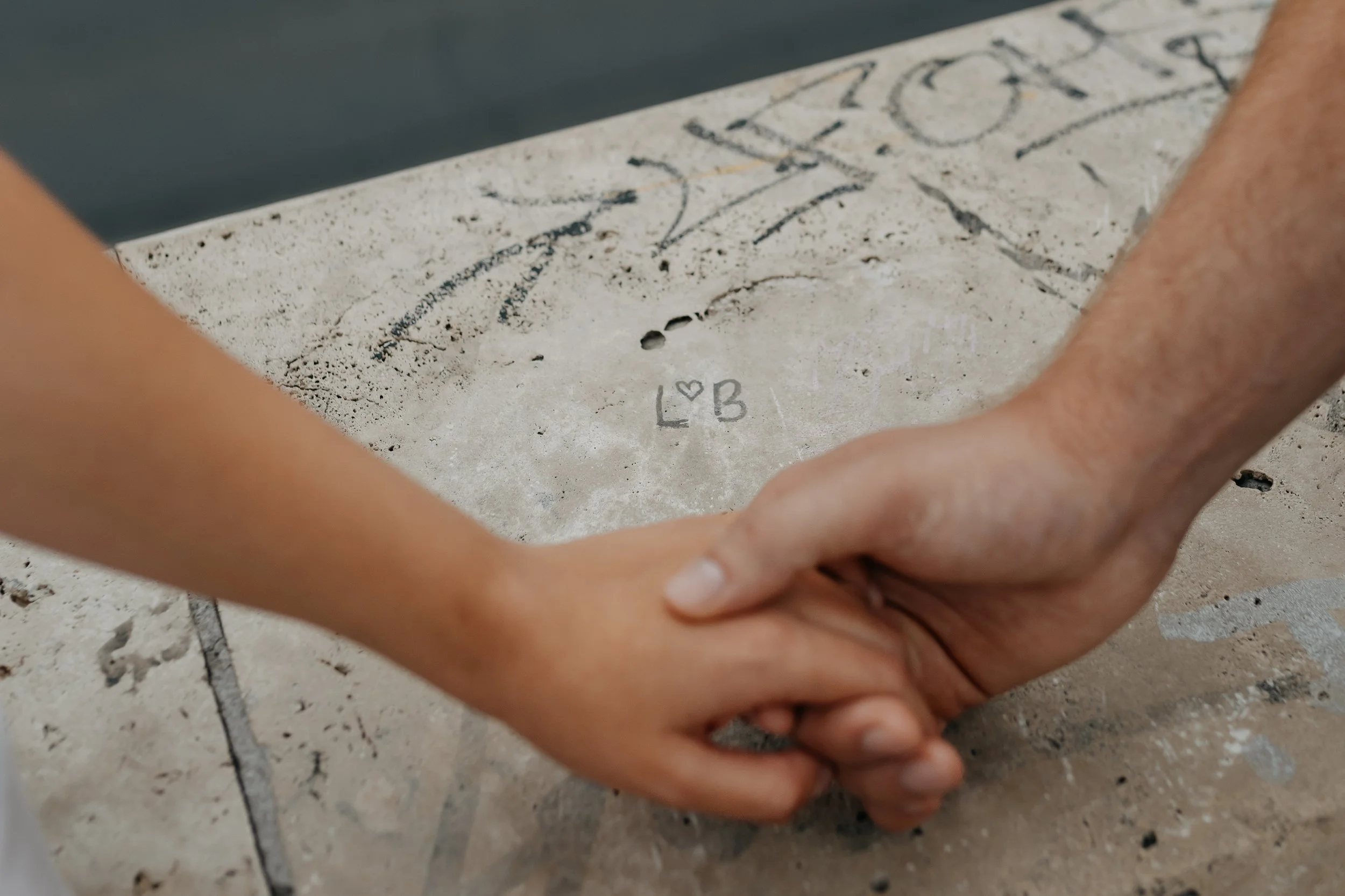 Two pairs of hands, one larger and one smaller, clasped together in an embrace on a tiled surface. The tiled surface has faint writing and drawings, including the letters 'B' and 'L' with a heart symbol.