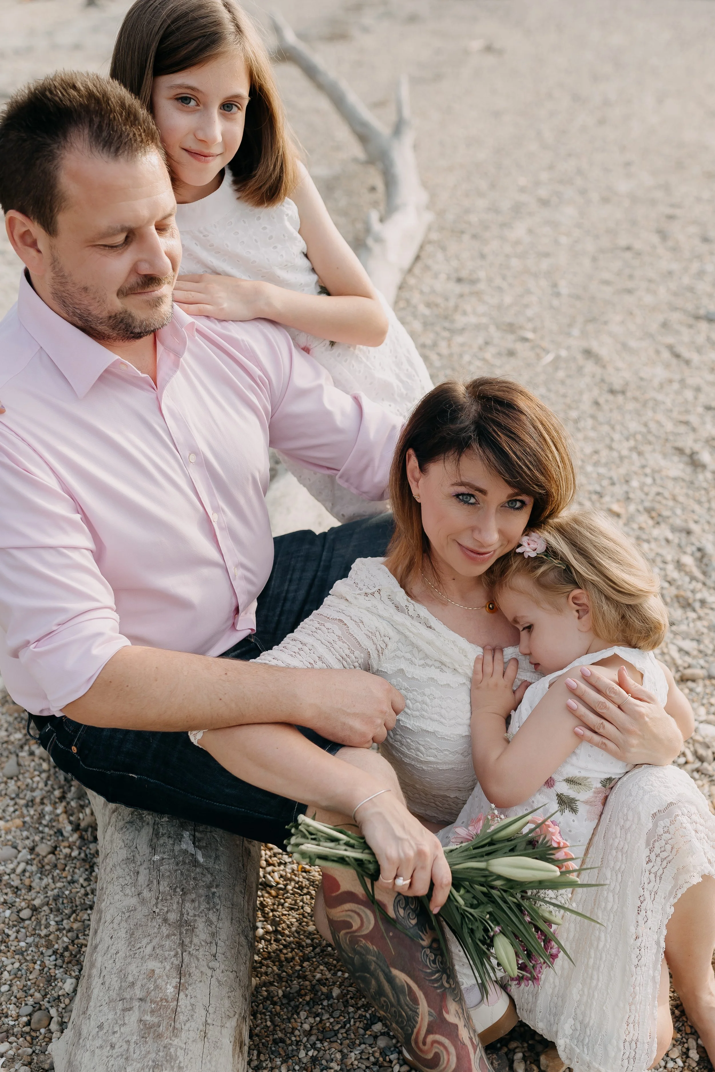 A family of four sitting on a cloudy day on a large log near a gravel area. The father, with a light pink shirt, has his eyes closed and is holding a bouquet of white flowers. The mother, with reddish-brown hair, is holding a young girl with blonde h