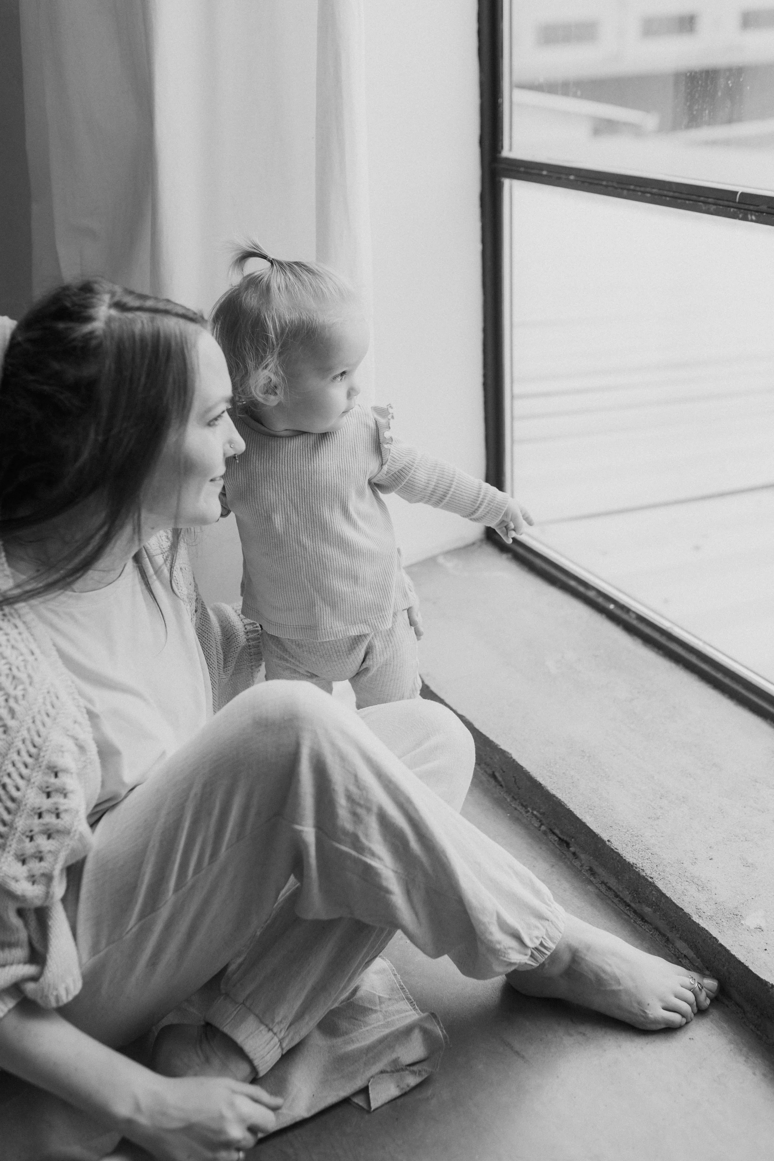 A woman and a young girl sitting on the floor near a large window, looking outside. The girl points at something outside the window.