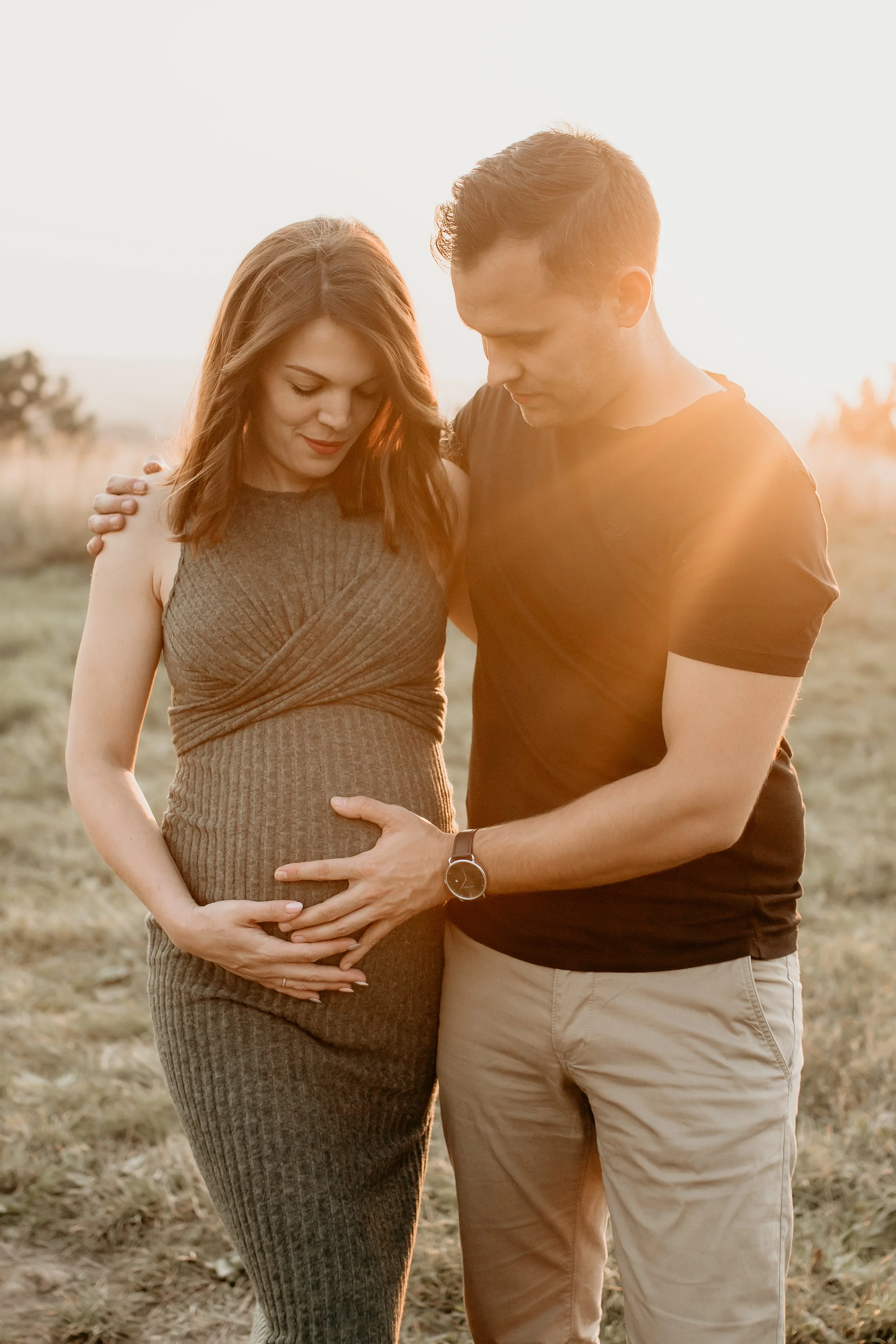 A pregnant woman and her partner standing close outdoors during sunset, with both hands on her belly and their heads bowed together in a tender moment.