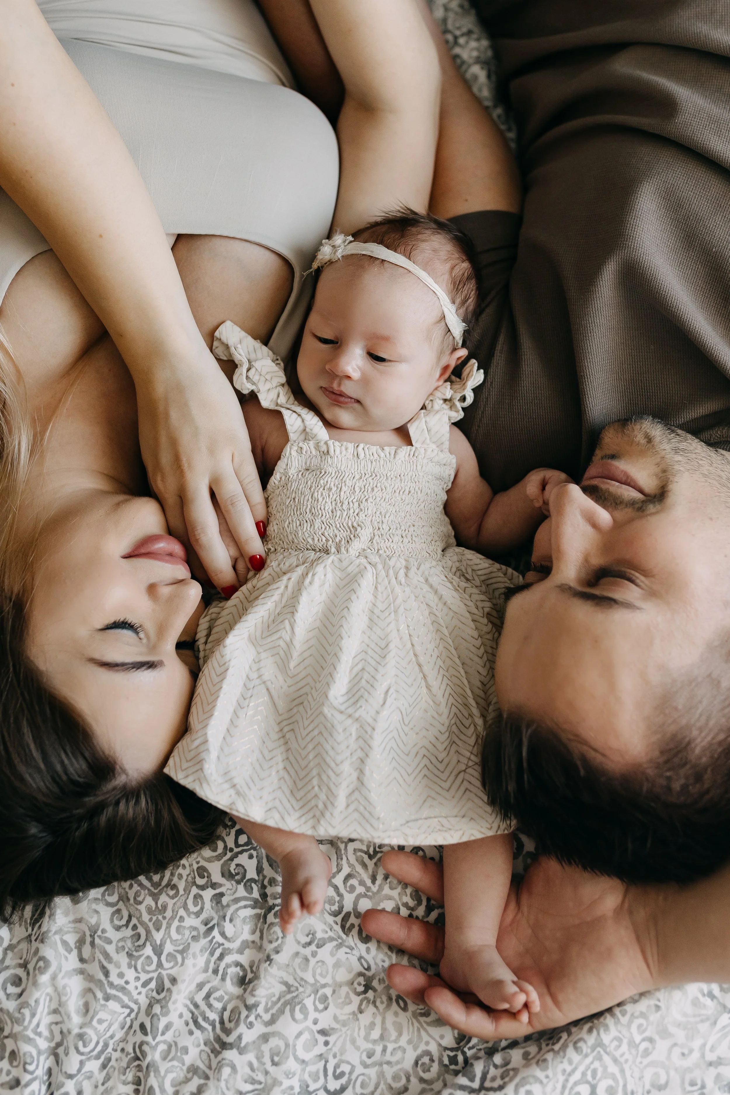 Family lying on a bed in a close-up, top-down view, holding hands and looking at each other, with a baby girl in the center.