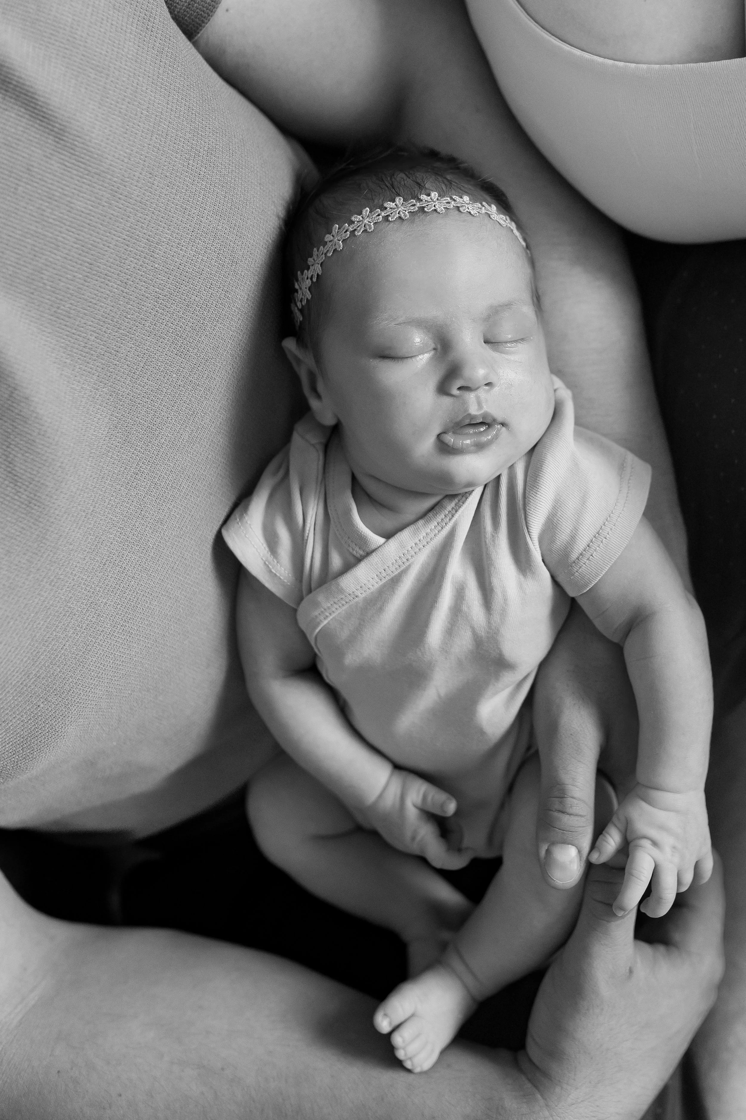 A black and white photo of a sleeping infant with a floral headband, being held gently by an adult. The baby's eyes are closed, and their mouth is slightly open.