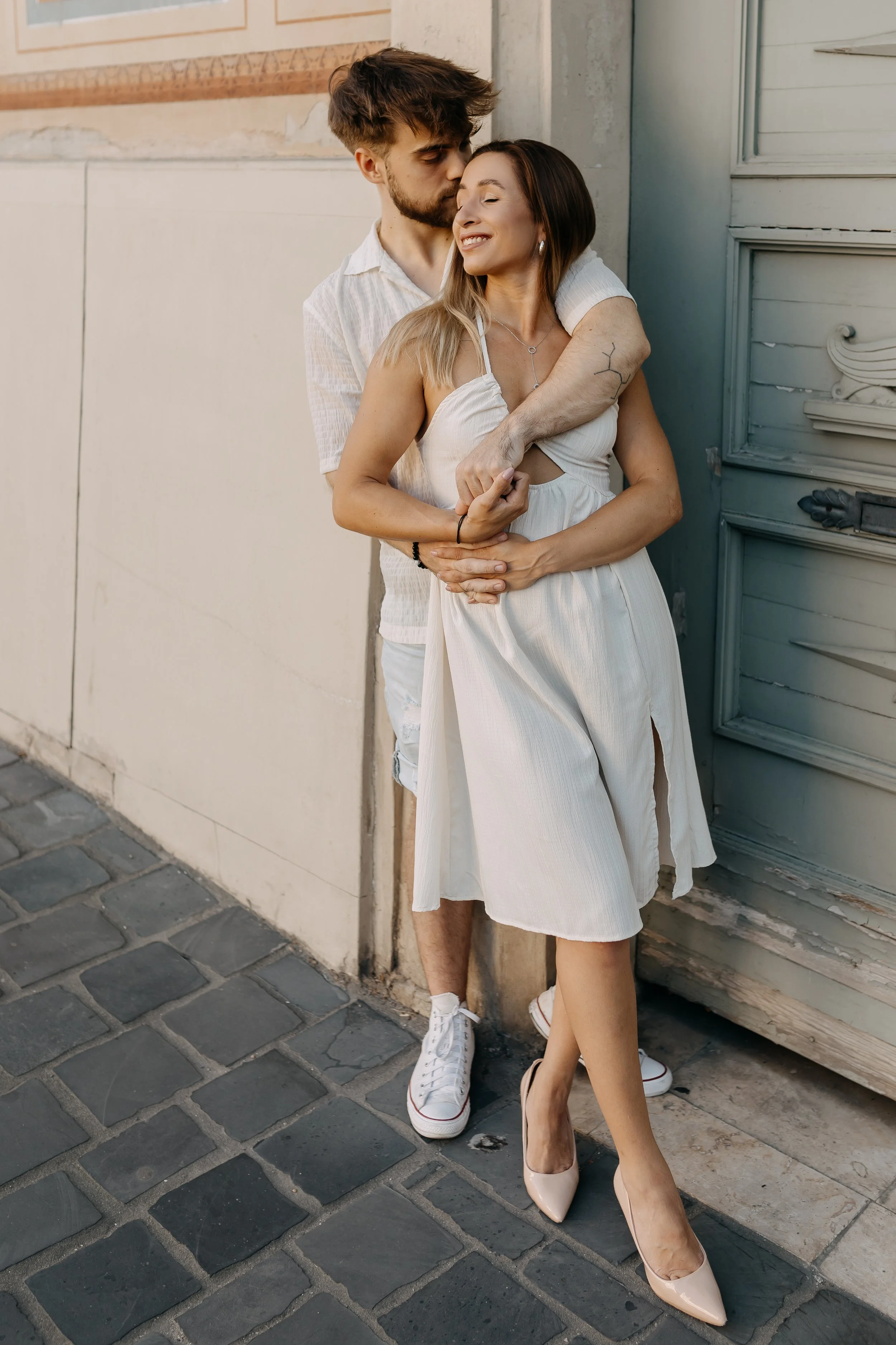 A young couple standing close together against a wall outside, with the man kissing the woman on her head while she smiles with her eyes closed. The woman is wearing a white dress and heels, and the man is in a casual shirt and shorts, both dressed i