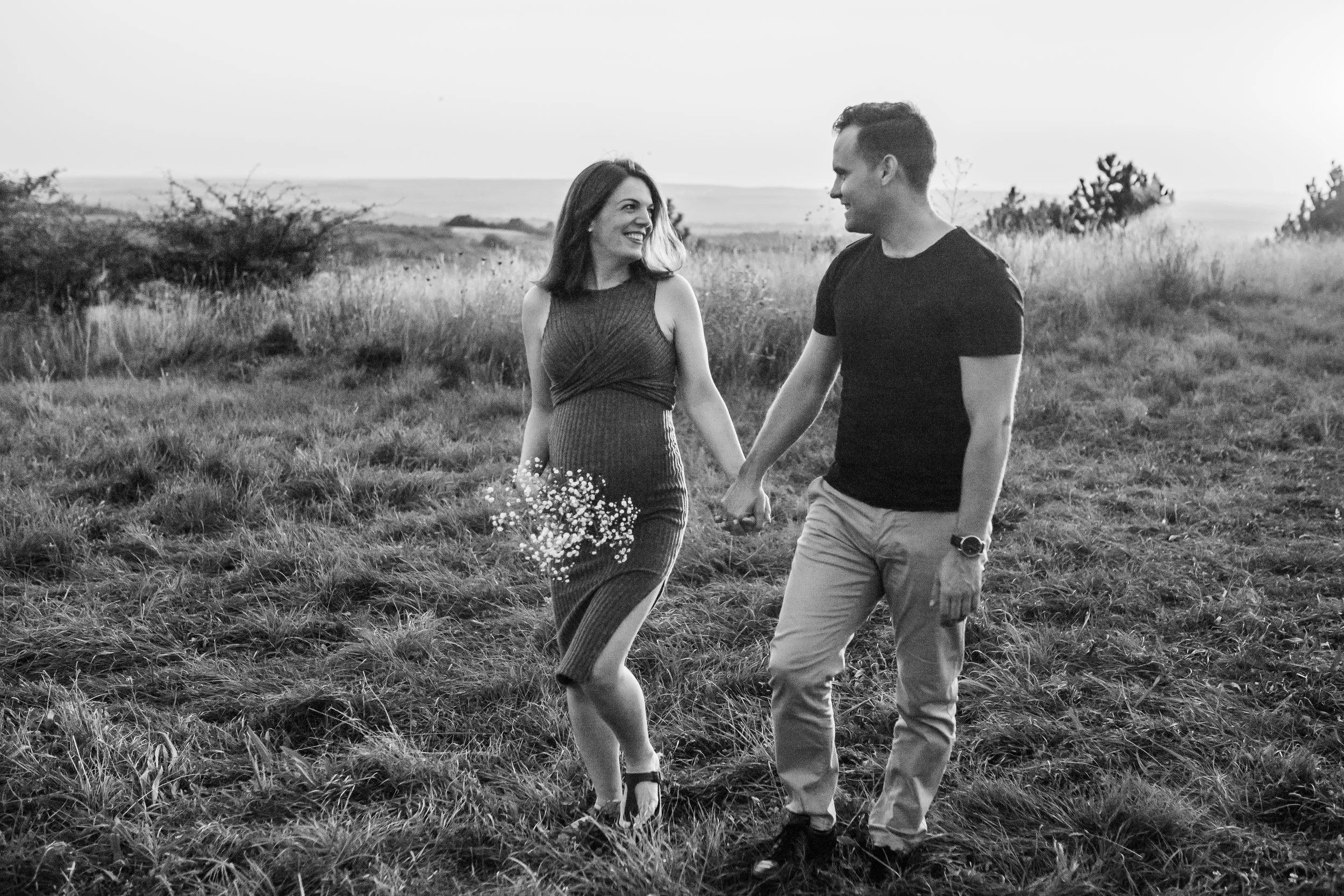 A black-and-white photo of a couple holding hands and smiling at each other, walking through a grassy field.