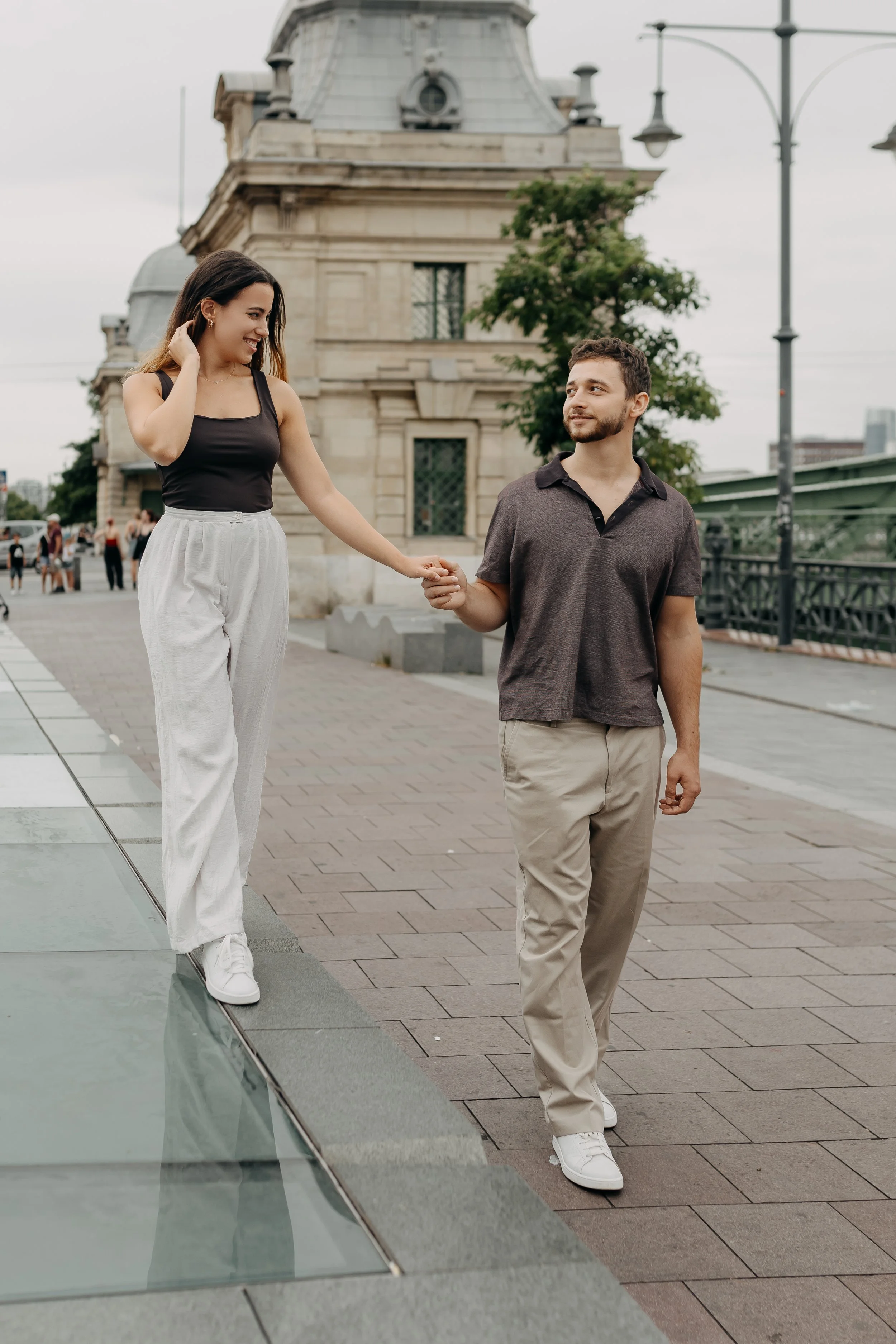 A woman in a black tank top and white pants smiling and holding hands with a man in a dark gray shirt and beige pants on a city street with a historic building and streetlamp in the background