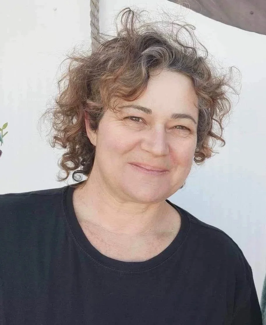 A woman with short curly hair and earrings, smiling, wearing a yellow and black safety vest and black shirt, standing inside a tent with other people around, holding a white bucket labeled 'Community Compost Nelson.'