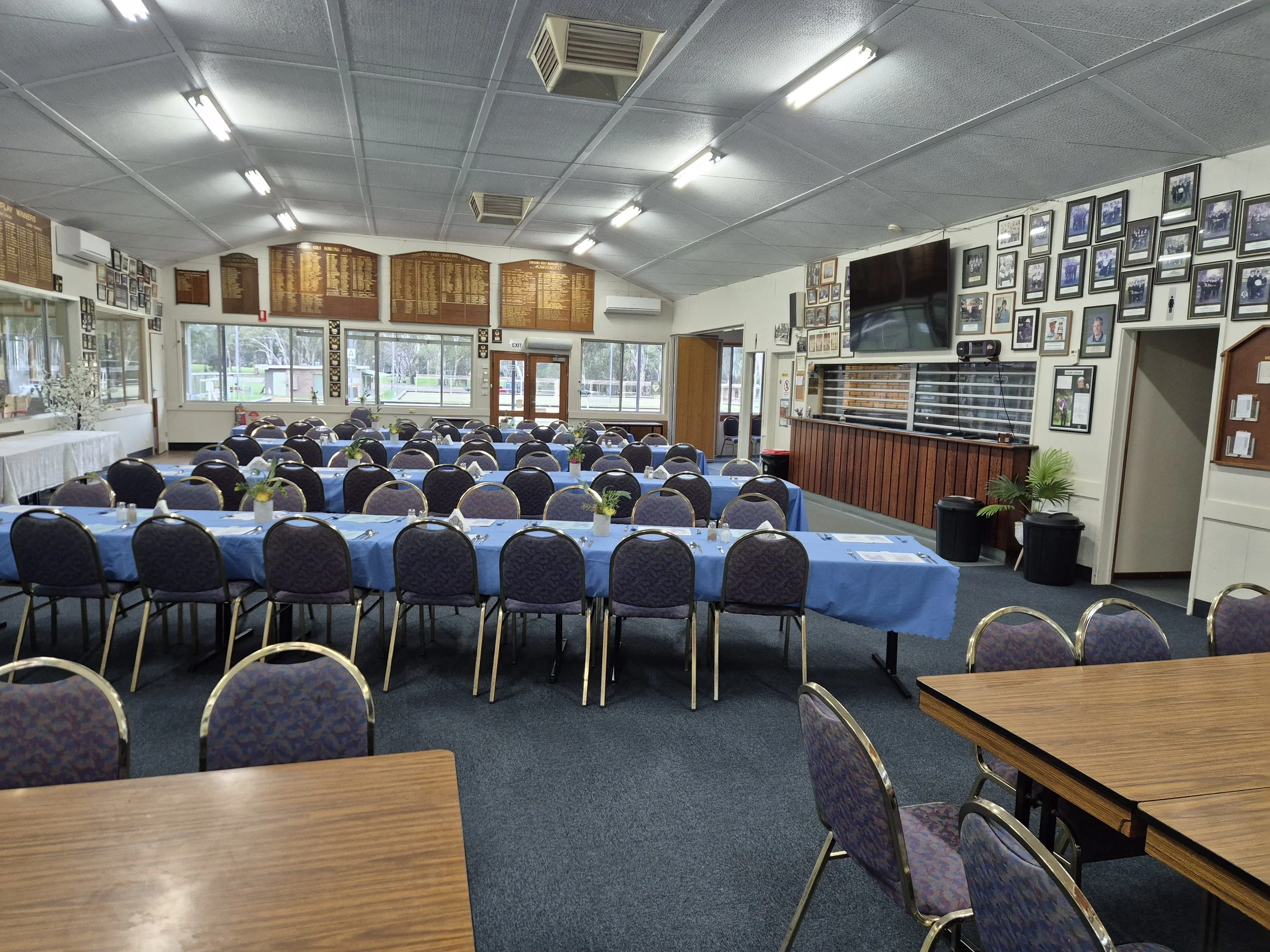This image shows the interior of a banquet hall or community room set up for an event, with rows of tables covered in blue tablecloths and surrounded by chairs, and a wall decorated with numerous framed photographs and plaques.