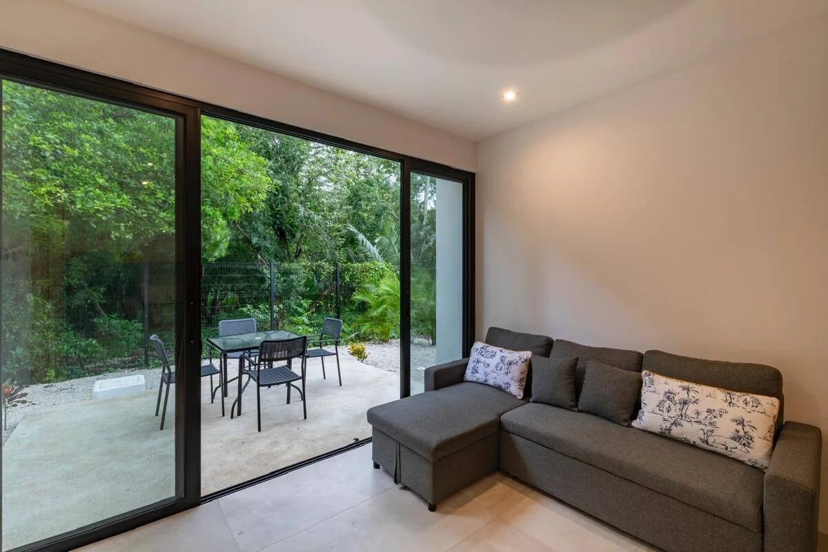 Living room with a gray sectional sofa with decorative throw pillows, large glass sliding doors leading to a patio with outdoor dining table and chairs, surrounded by lush green trees.