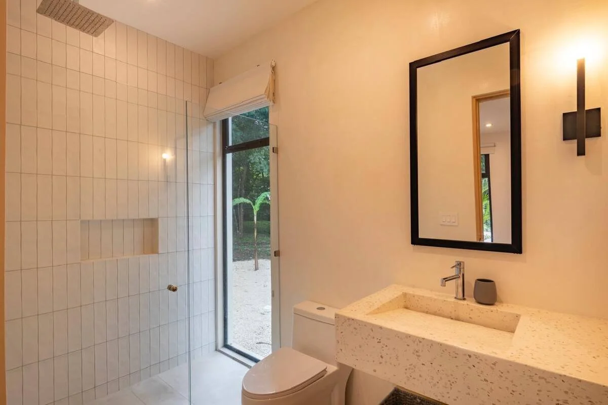 A modern bathroom with beige tiled walls, a walk-in shower, a white toilet, a vanity with a rectangular stone sink, a large black-framed mirror, and a wall sconce light, with a glass door leading to an outdoor garden.