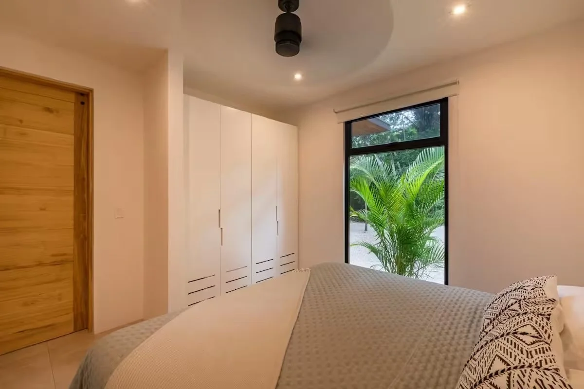 A bedroom with a closed wooden door, a large closet with white doors, a window with a view of tropical plants outside, and a bed with patterned pillows and a textured blanket.
