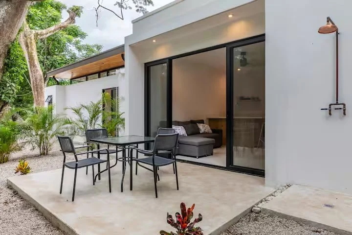 Outdoor patio with a round black patio table and four black chairs, adjacent to a modern house with large sliding glass doors, a gray couch visible inside, potted plants, and a wall-mounted outdoor shower, surrounded by trees and greenery.