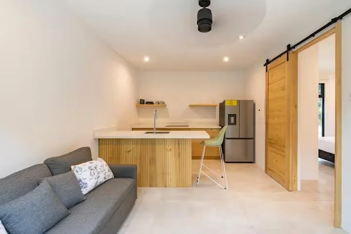 Modern kitchen and living space with a gray sofa, white walls, and wooden accents, including sliding barn doors and shelves.