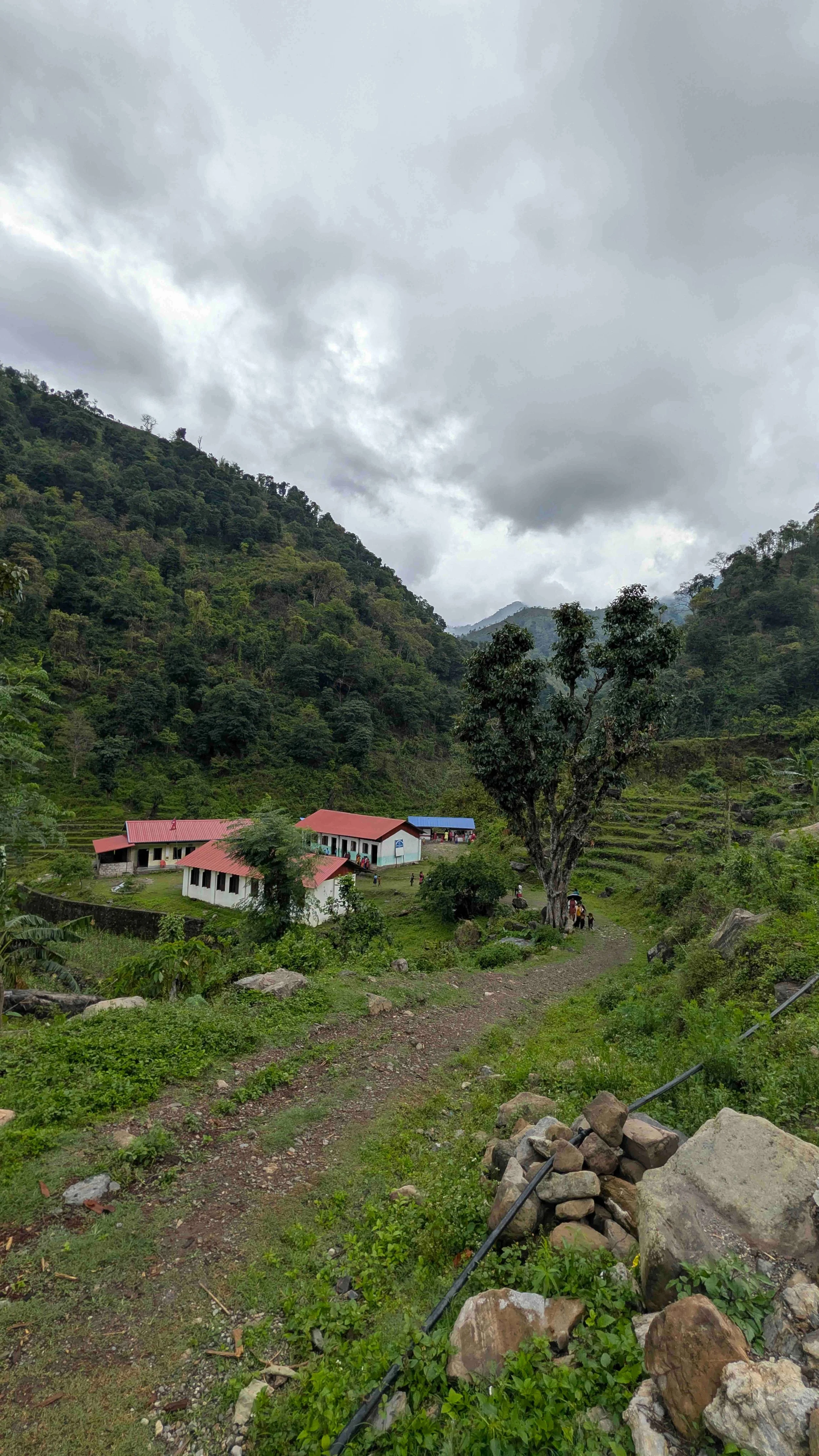 Ein Schulgelände mit roten Dächern in einem grünen Gebirgstal in Chepang, Nepal unter bewölktem Himmel