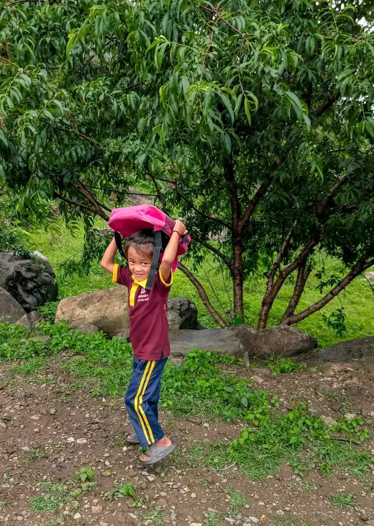 Ein lächelndes Mädchen in weinroter Schuluniform hält seine pinke Schultasche über den Kopf, an den Füßen Schlappen, auf dem steinigen Weg an der Bergschule in Chepang, Chitwan, Nepal