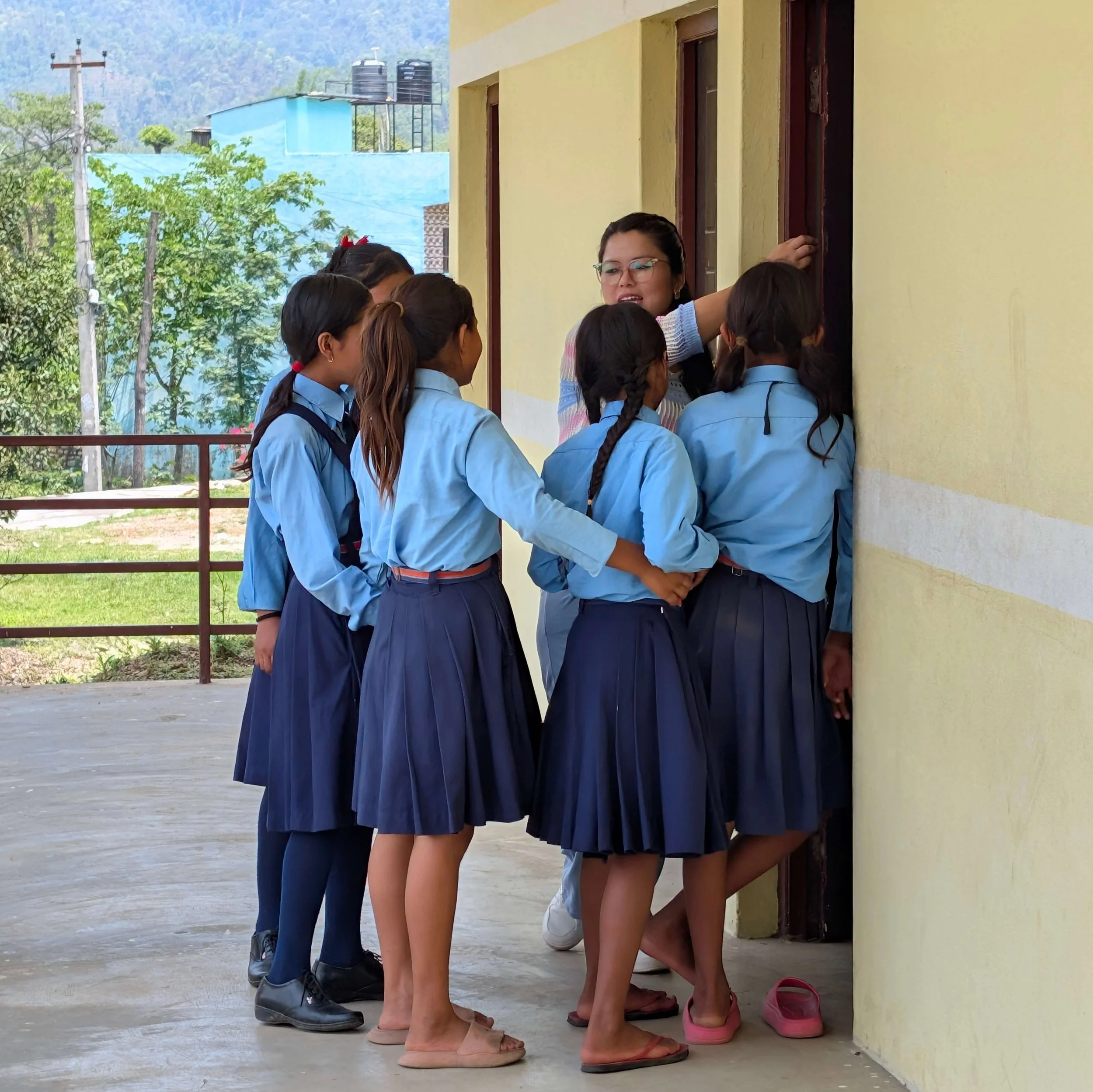Eine Gruppe Schülerinnen in hellblauen Uniformen und dunkelblauen Faltenröcken steht vor einem Klassenzimmer der Schule in Thimura bei Bharatpur, Nepal, im Gespräch mit der Lehrerin