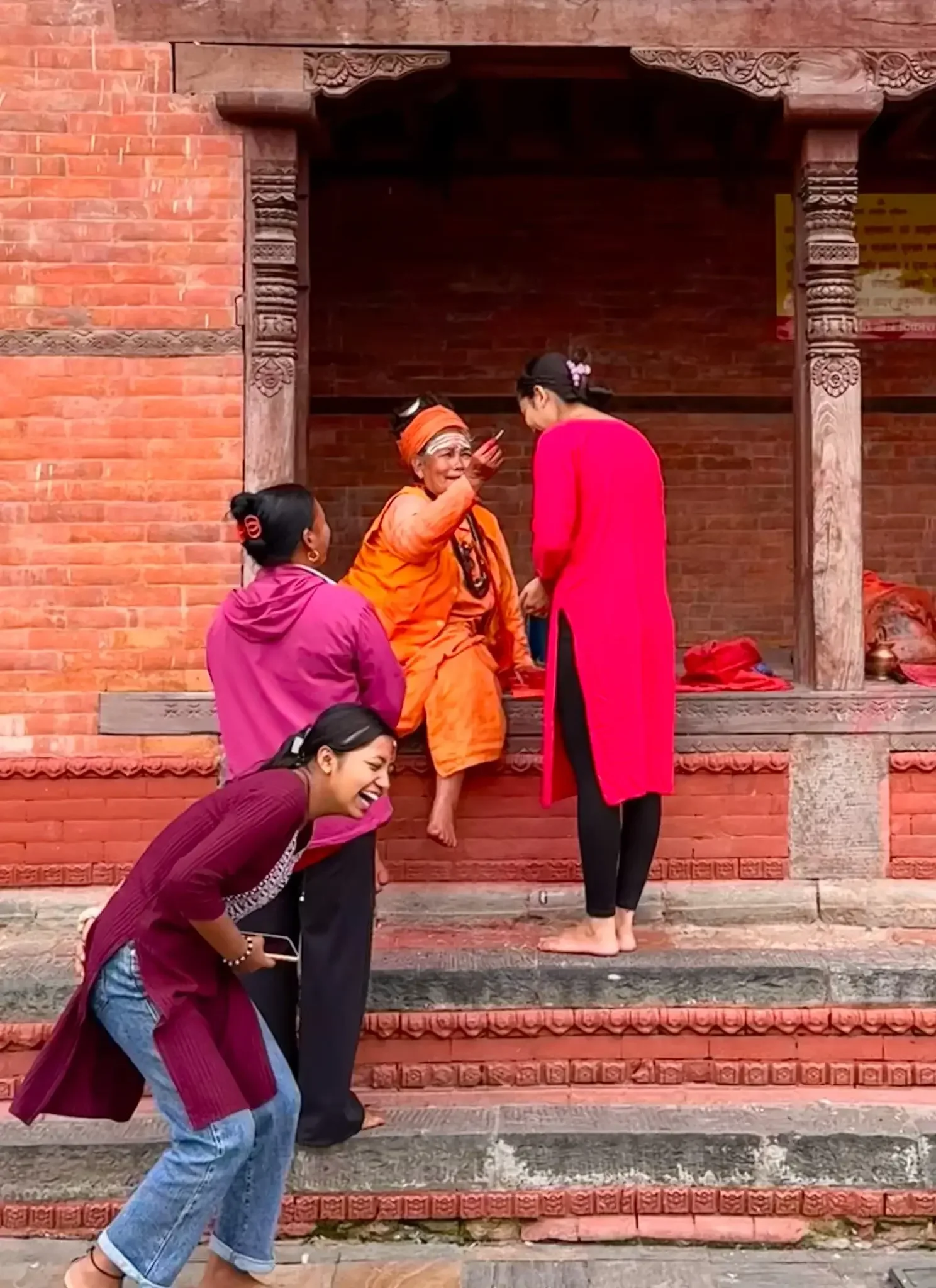 Junge Frauen interagieren freudig mit einer älteren Frau in orangefarbenem Gewand vor einem Gebäude mit traditionellen Holzschnitzereien in Nepal