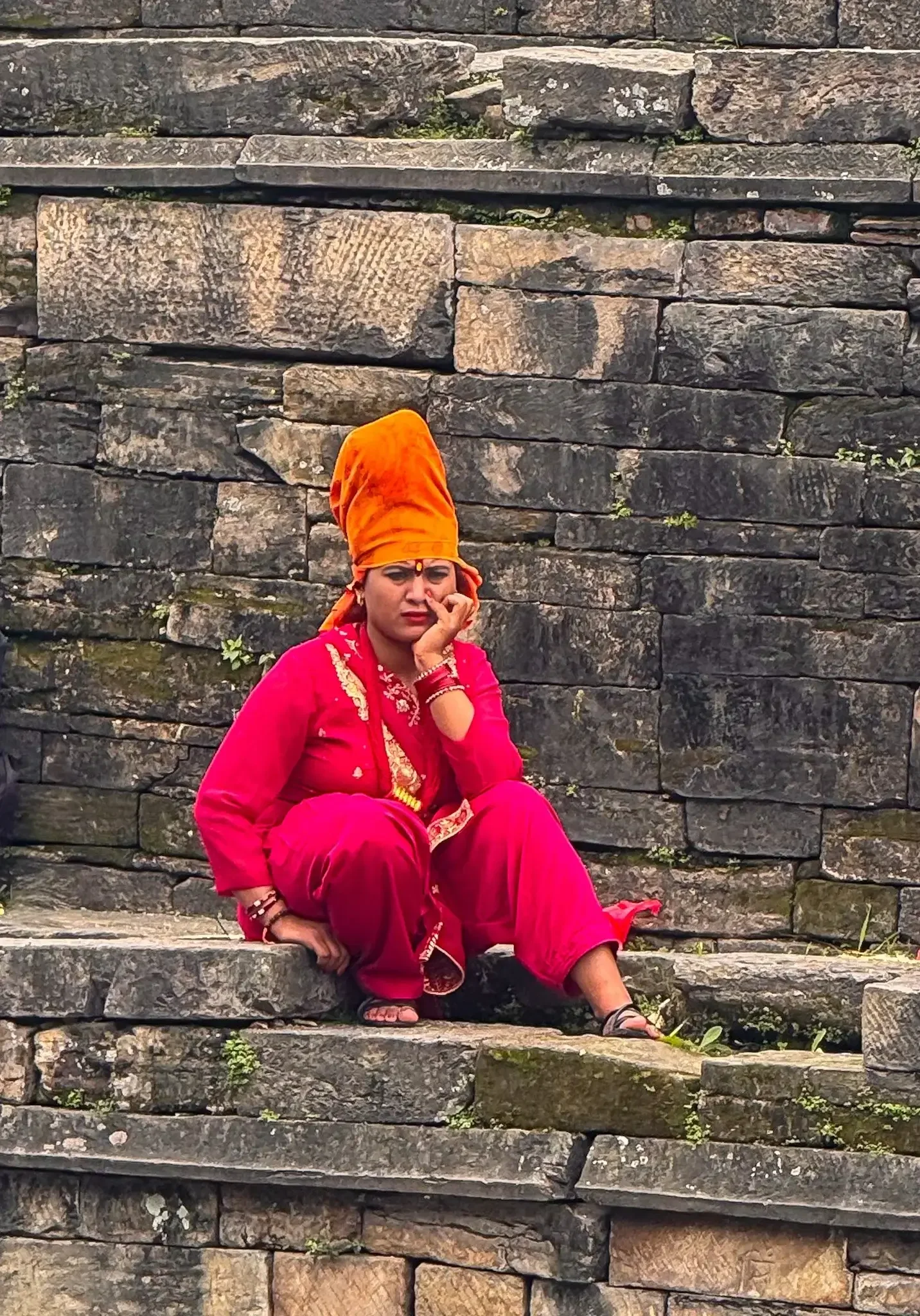Eine Frau in leuchtend roter Kleidung und orangefarbenem Turban sitzt auf steinernen Stufen in Nepal und blickt nachdenklich zur Seite