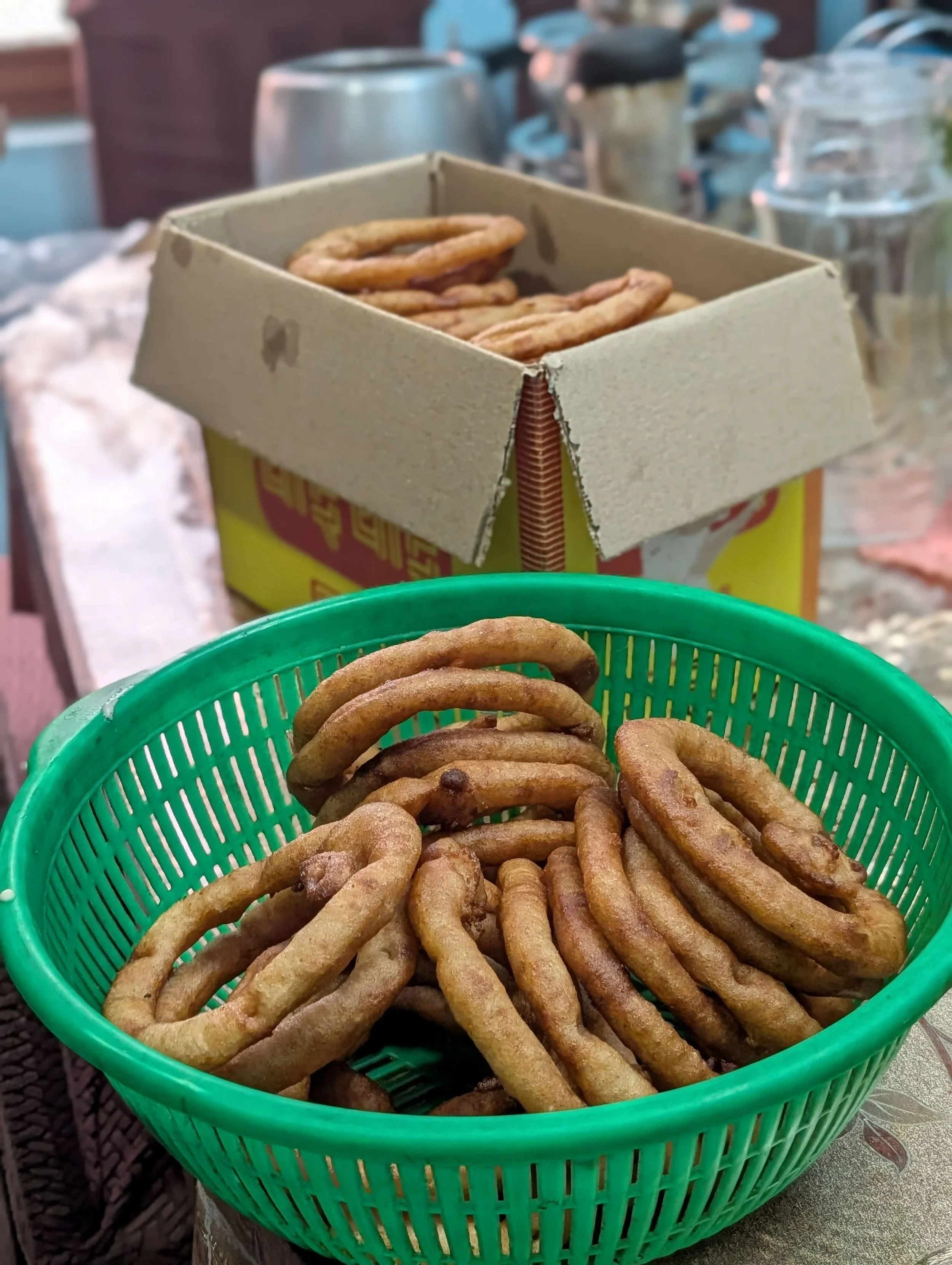 Mehrere ringförmige Sel Roti liegen in einem grünen Sieb und dahinter in einemgeöffneten Karton.
