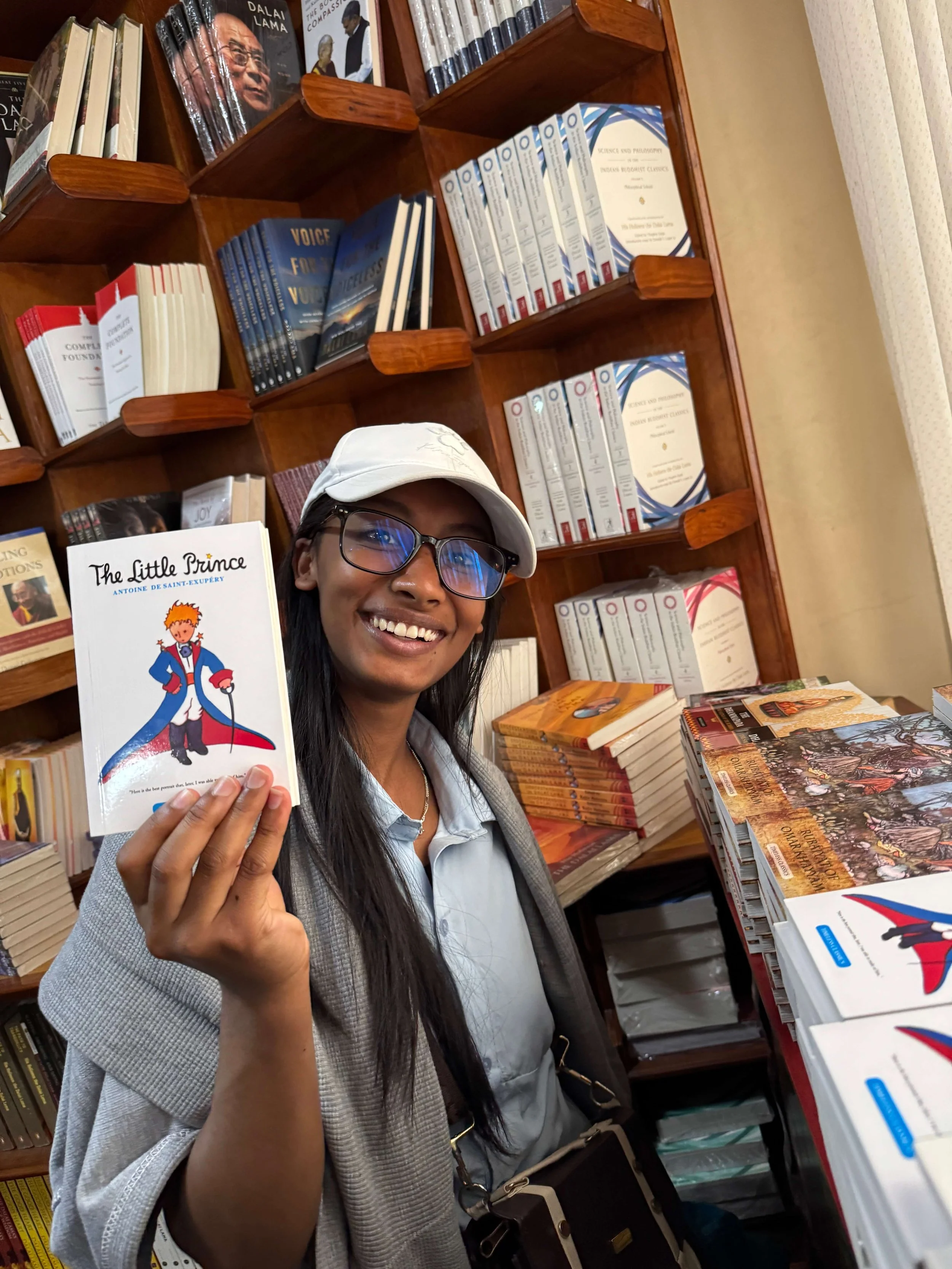 Eine junge Frau mit weißer Cap und Brille lächelt in der Buchhandlung des Kopan Klosters und hält die englische Ausgabe von „The Little Prince" in die Kamera, hinter ihr Regale mit buddhistischer Literatur.